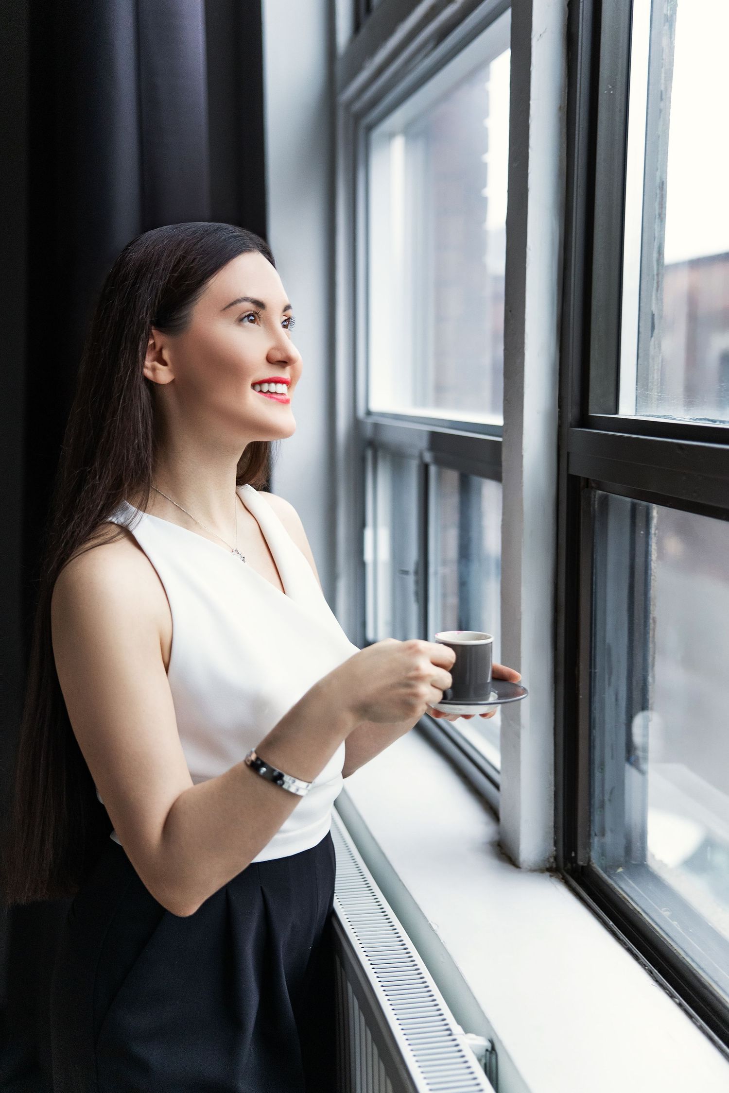 Smiling brunette woman holding coffee cup looking out large window in modern interior setting.