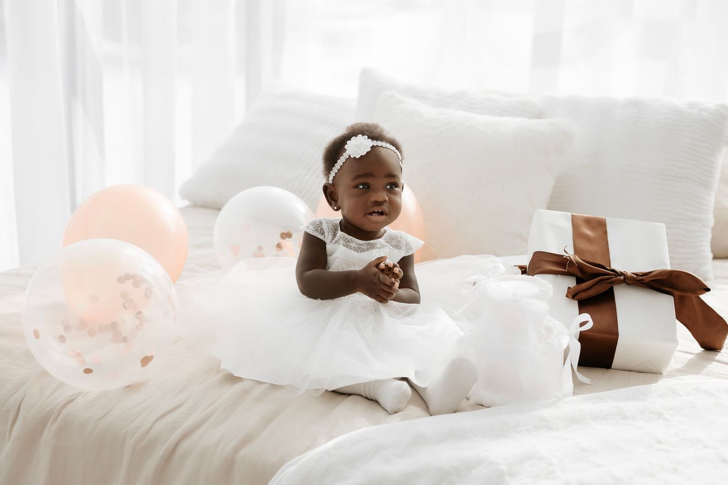 Adorable baby girl in white dress sitting on bed with balloons and gift box at first birthday celebration.