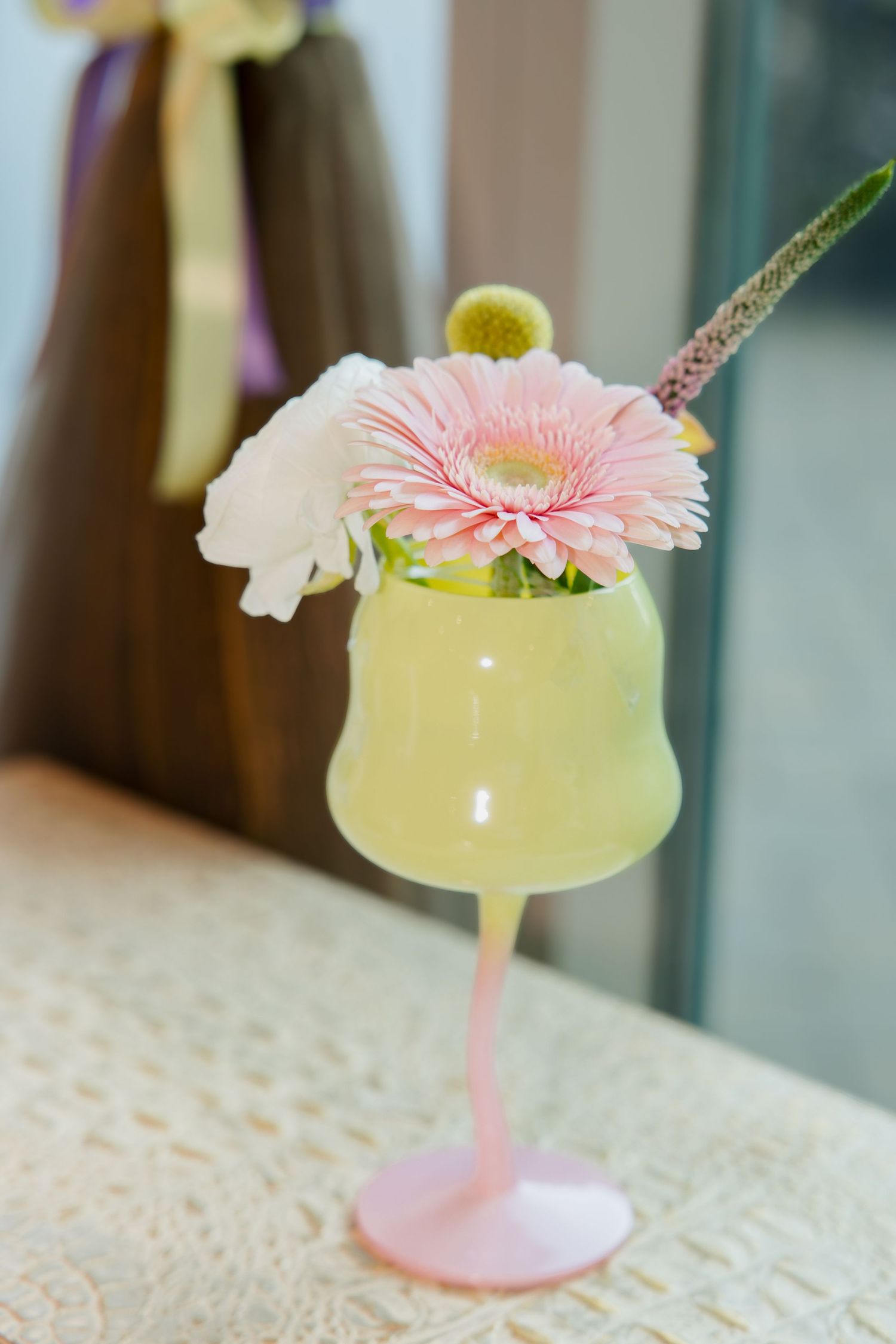 A yellow cocktail in a pink stemmed glass garnished with a pink gerbera daisy, white flower, and lime slice.