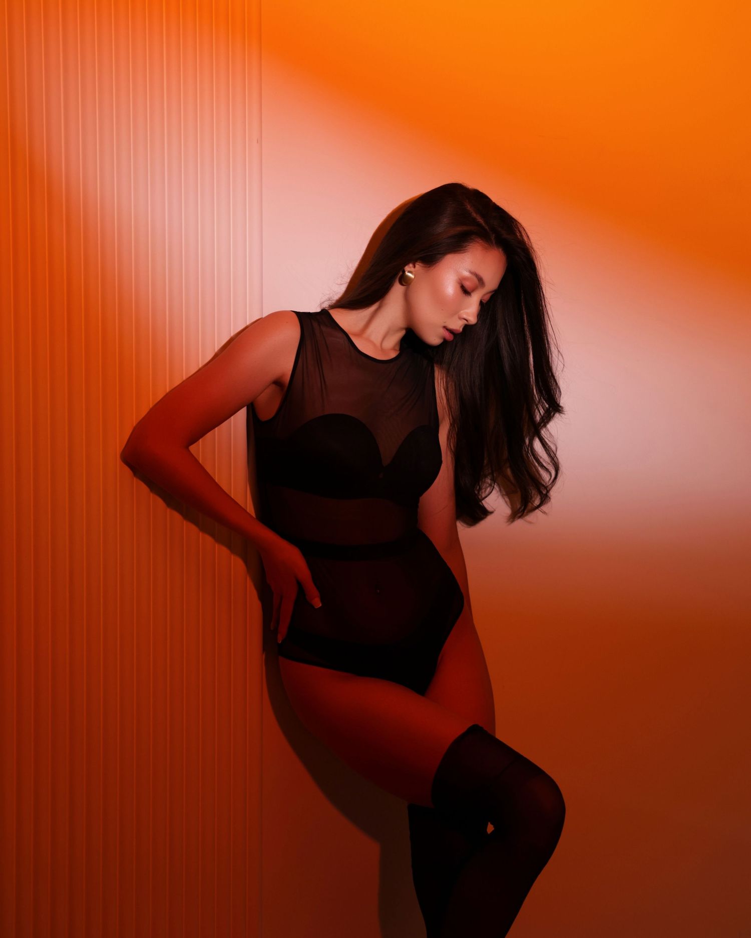 Asian woman in a black sheer bodysuit posing against a vibrant orange illuminated striped wall in a studio.