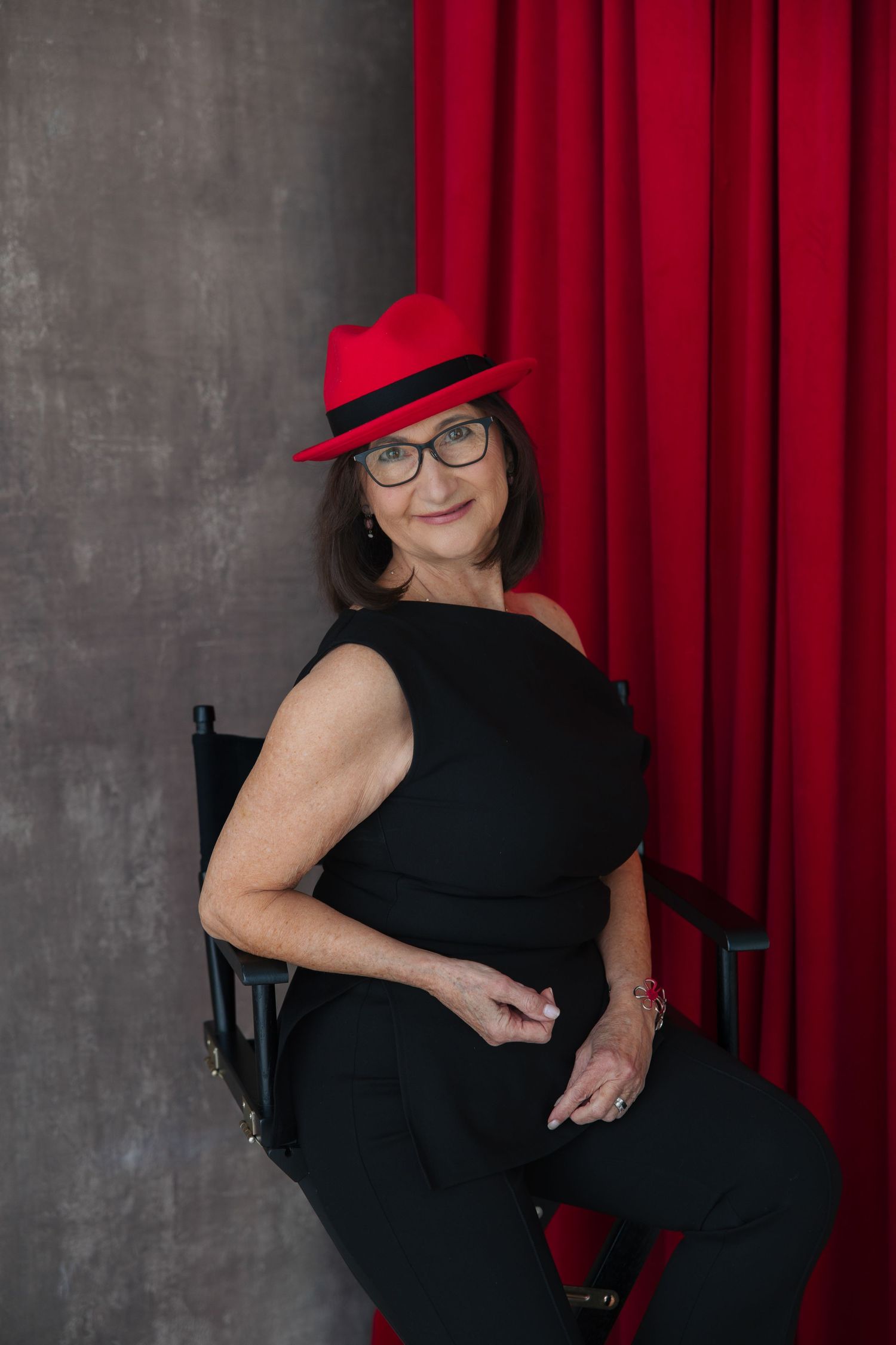 Stylish woman in black outfit and red fedora hat poses confidently in a director's chair.