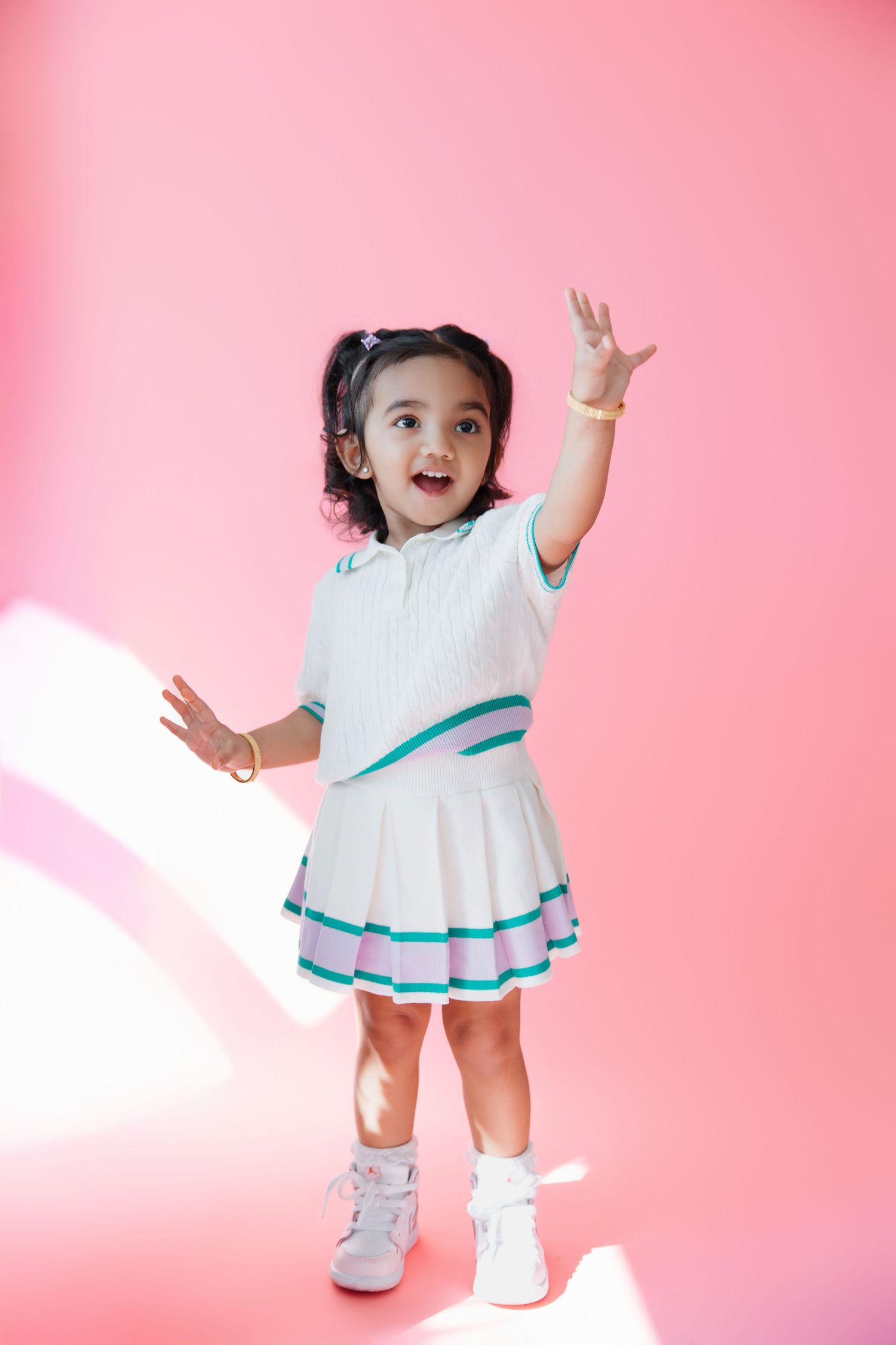 Joyful toddler girl in white tennis dress with teal trim, arms raised, posing on pink background.
