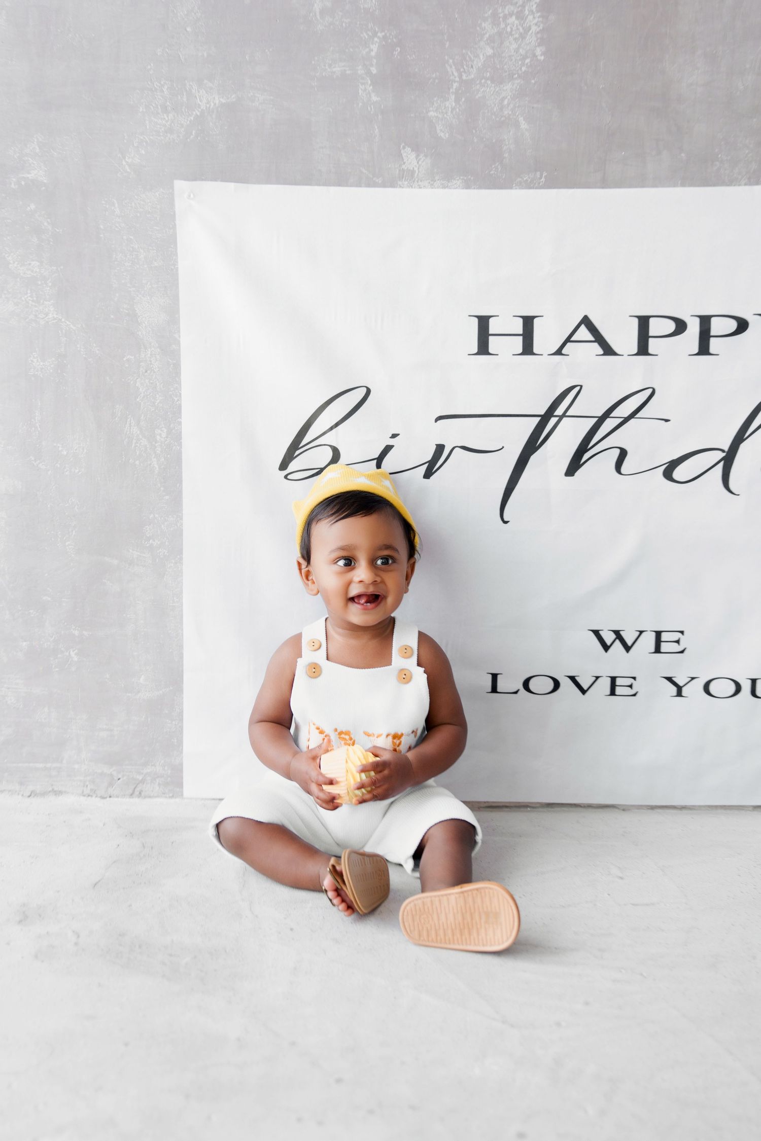 A smiling baby in white overalls and yellow crown hat sits before a Happy Birthday banner holding a glowing candle.