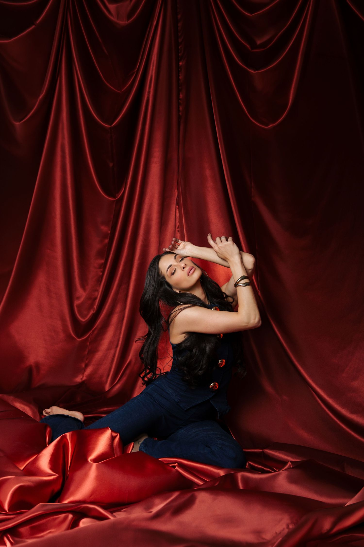 Brunette woman in navy outfit posing dramatically on red satin fabric against rich velvet curtains.