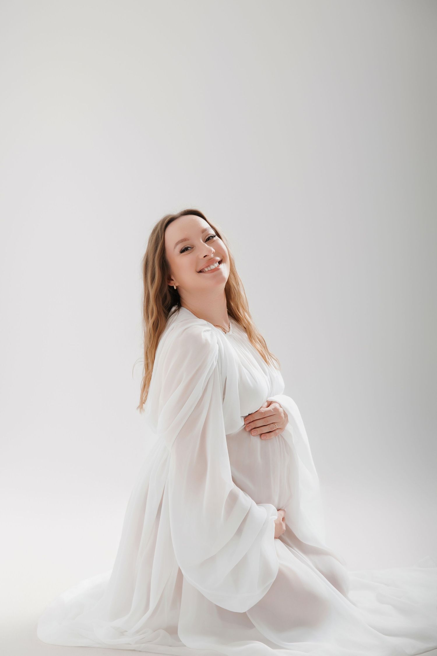 Smiling pregnant woman in flowing white dress sitting gracefully on floor against light background.