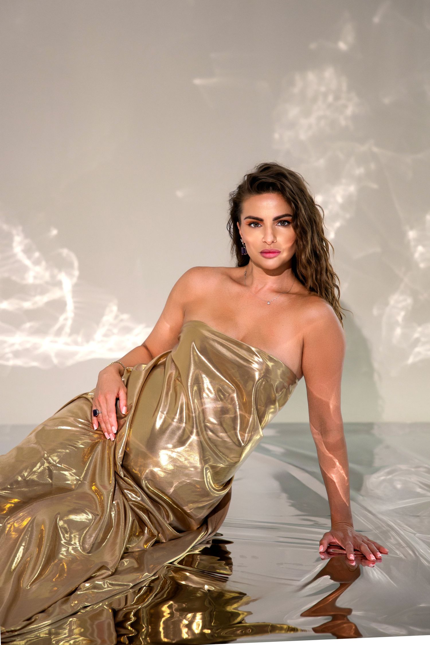 Woman in a gold metallic strapless gown posing elegantly in shallow water against a light gray background.