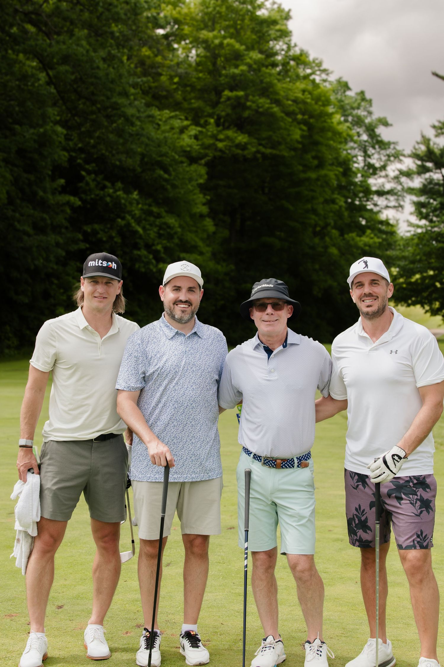 Four men in golf attire smile together on a lush green golf course on a sunny day.