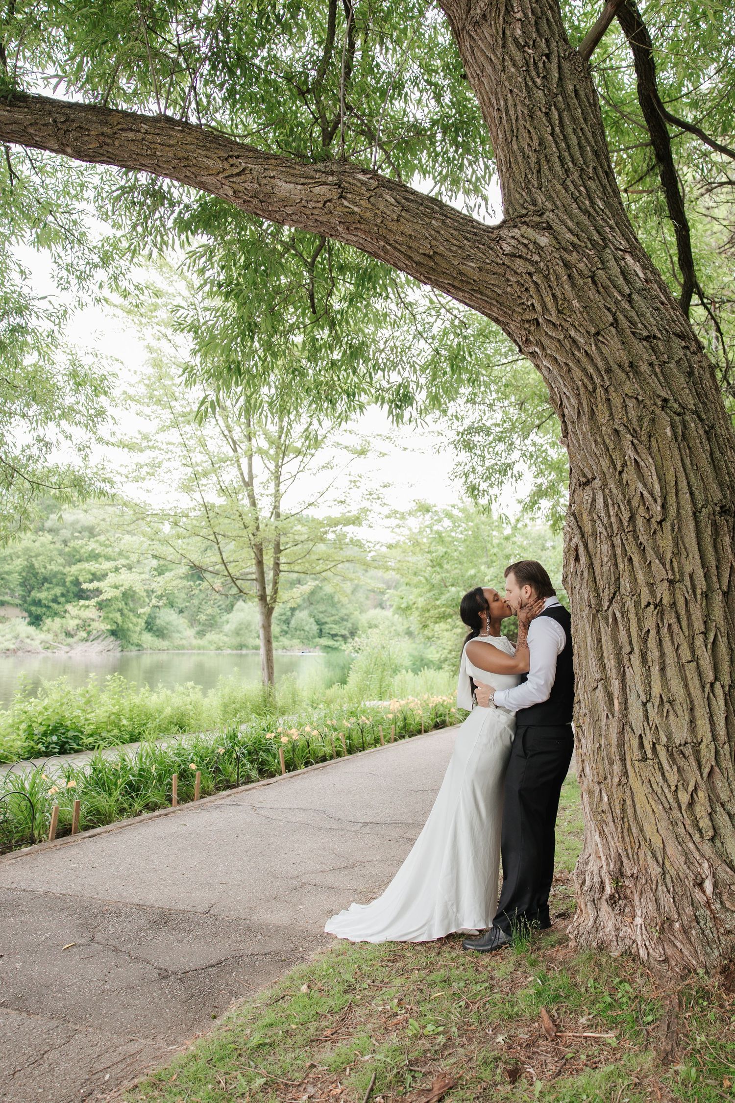Bride and groom kissing under a large tree by a serene pond, surrounded by lush greenery on their wedding day.