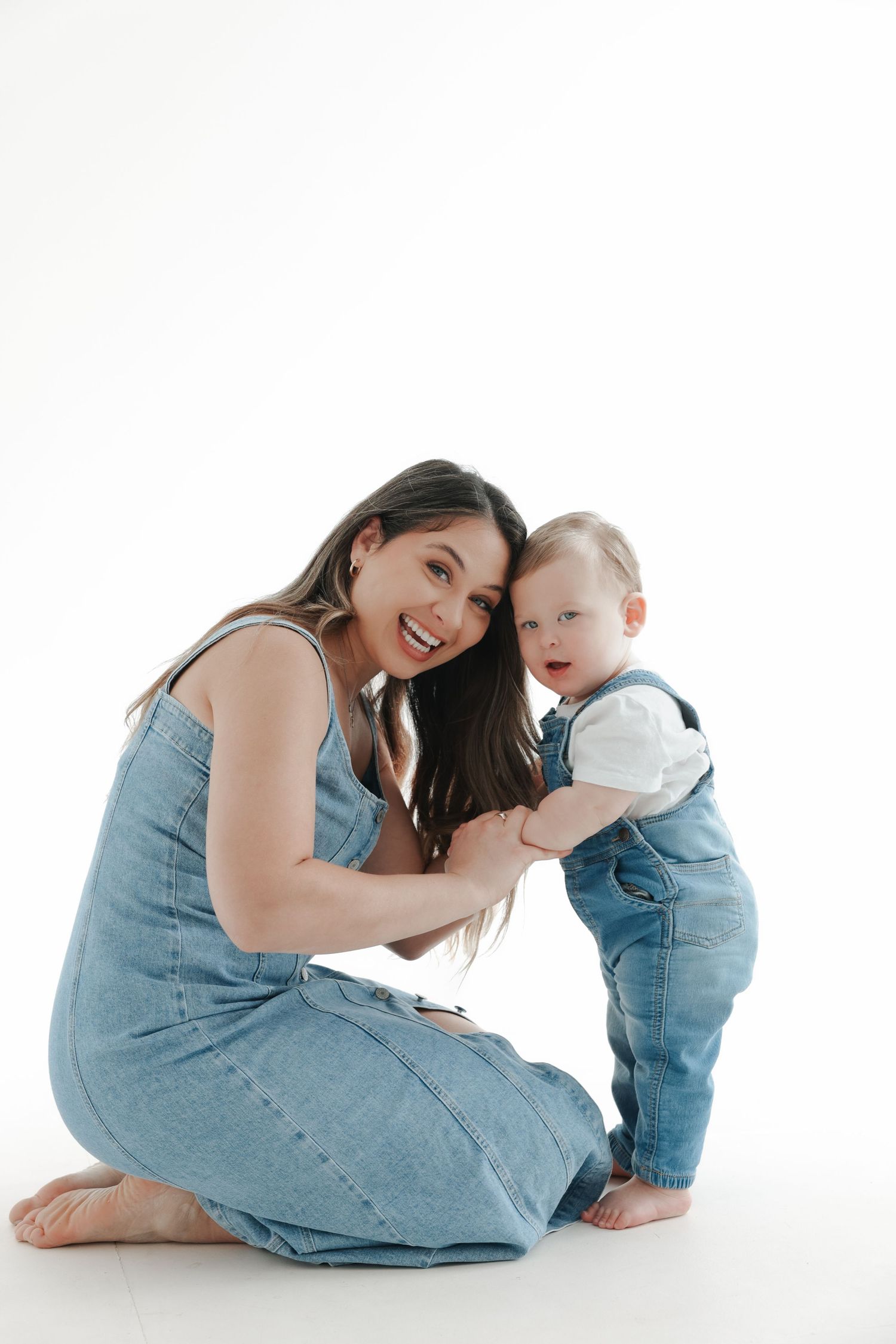 Smiling mother in denim dress sits with baby in denim overalls on white background.