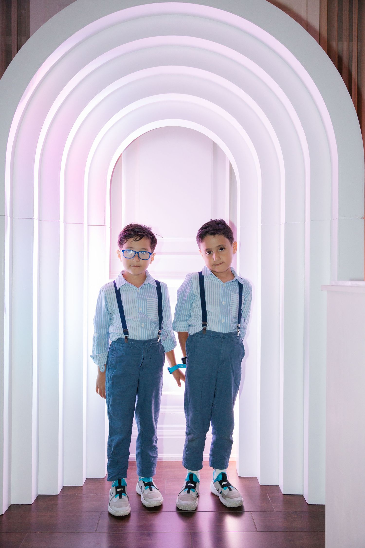 Two twin boys in matching blue shirts, suspenders, and jeans pose under white arched hallway with soft purple lighting.