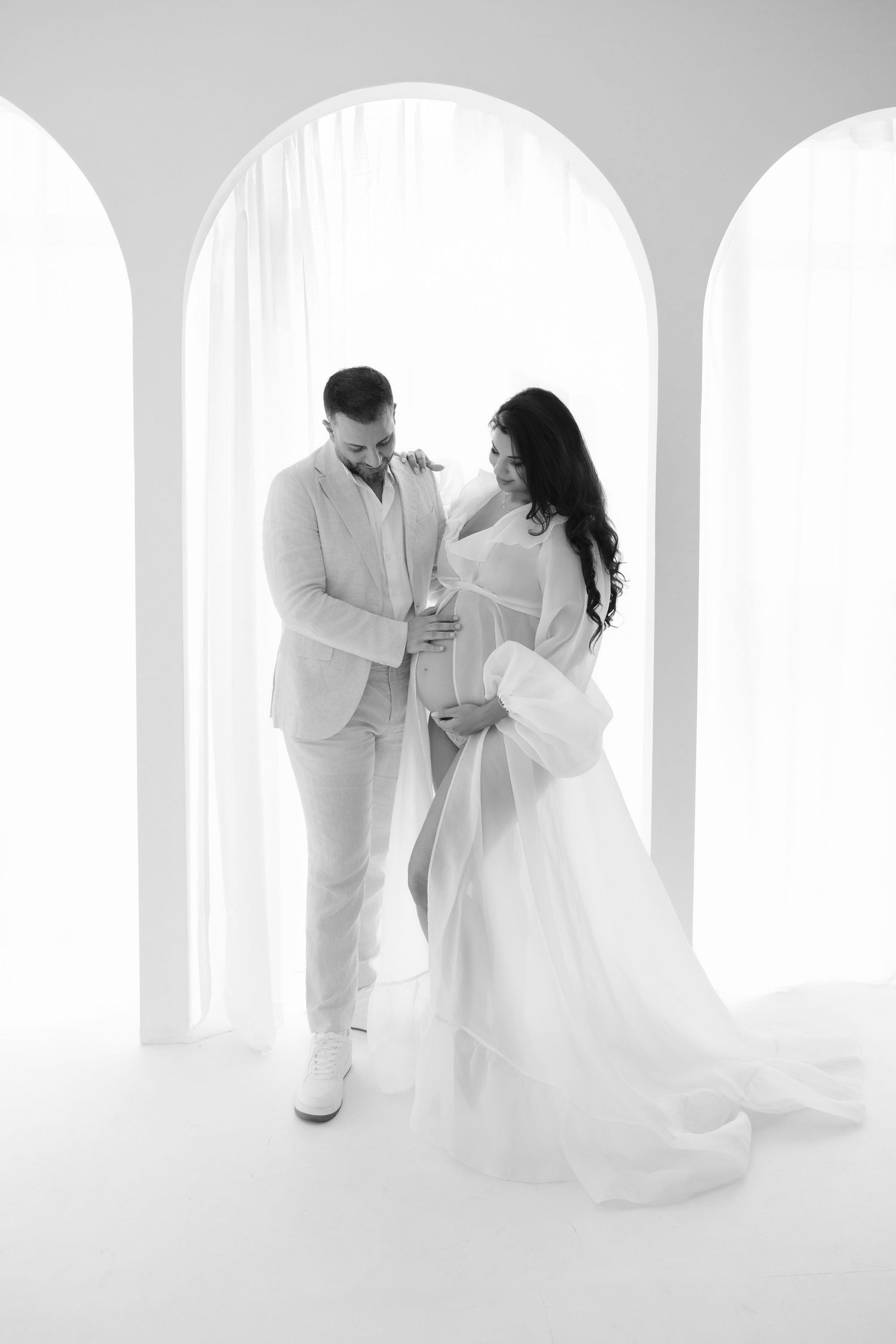 Black and white photo of a couple tenderly cradling a pregnant belly in an elegant white studio setting.