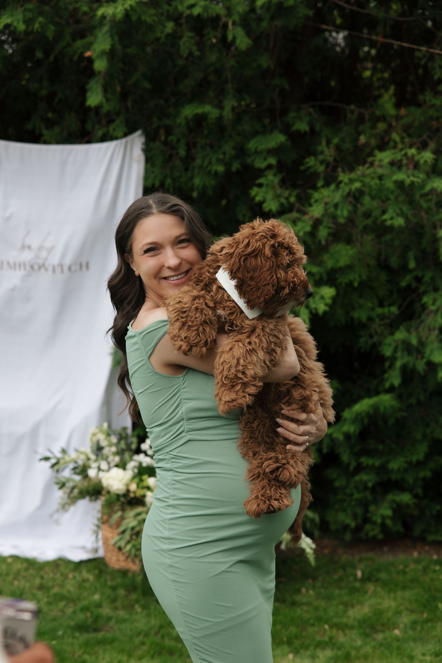 Smiling woman in green dress holding a fluffy brown doodle dog at an outdoor event.