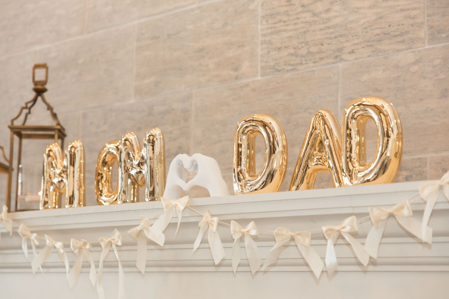 Gold foil MON and DAD letter balloons on a white mantel with cream ribbon bows and a lantern.