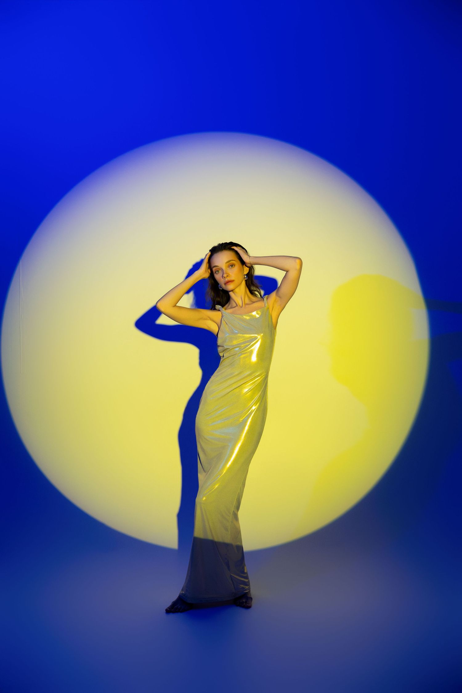 Woman in silver metallic gown posing before circular yellow light on blue background.