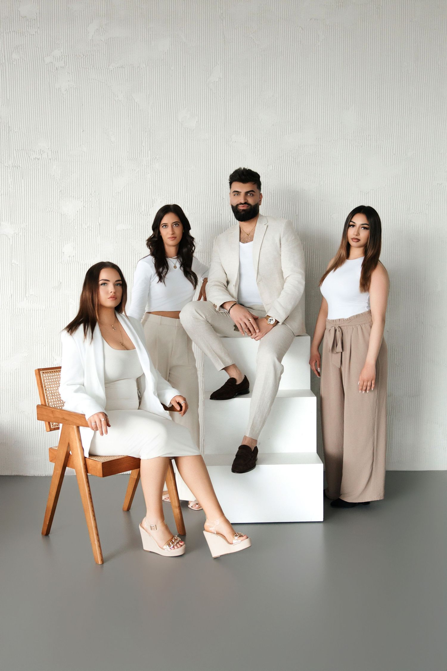 Four models in elegant white and beige outfits posing on white steps and chair in minimalist studio setting.