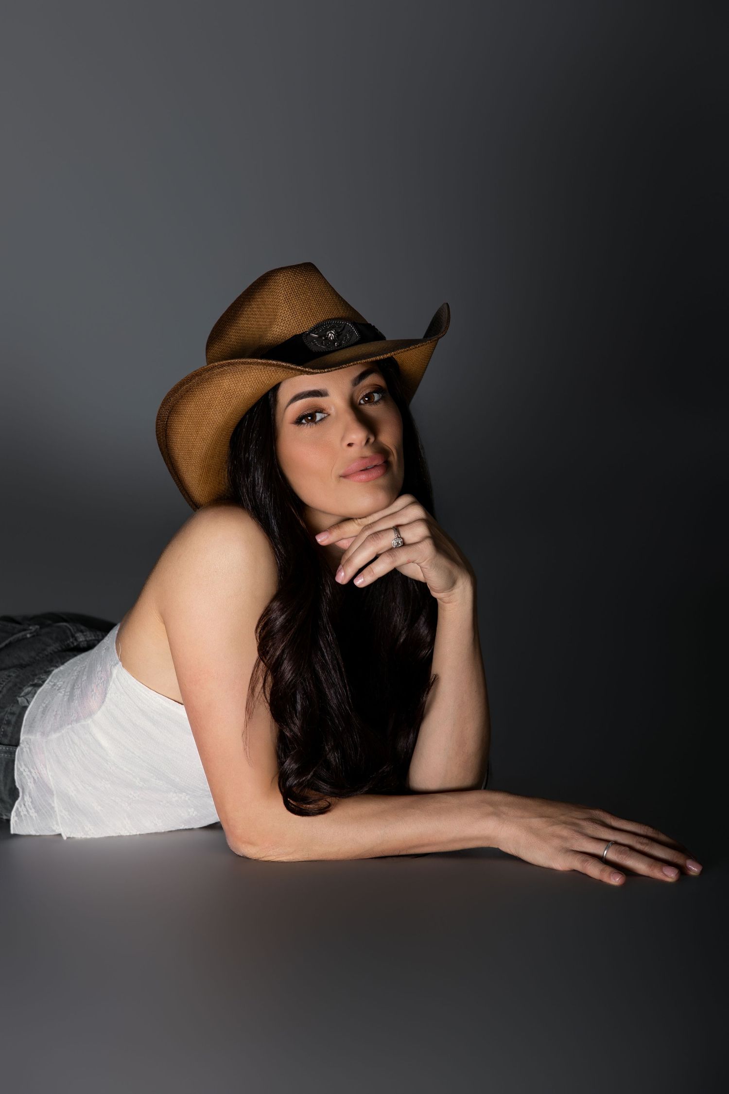 Brunette woman in white top wearing brown cowboy hat posing thoughtfully against dark gray background.