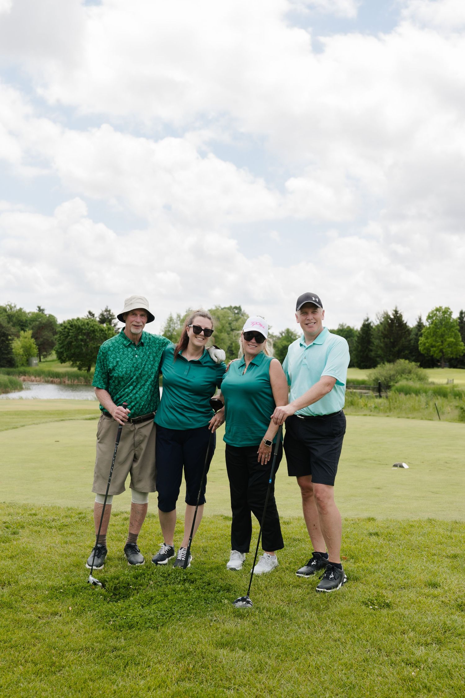 Four golfers in green shirts posing together on a lush golf course under a cloudy sky.
