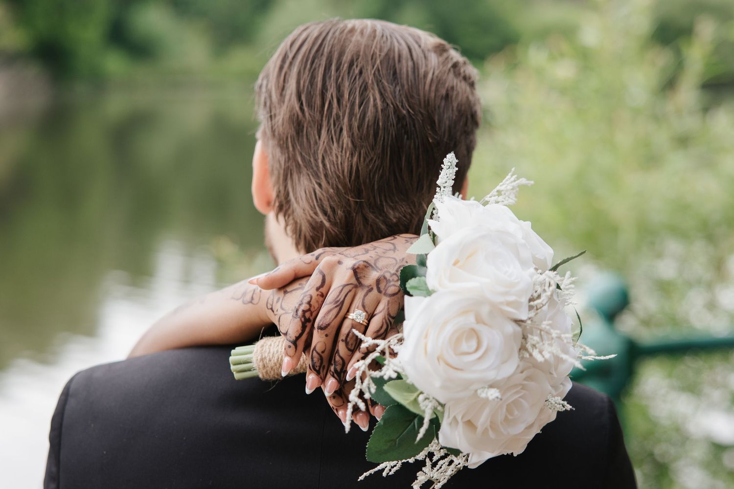 Bride resting her hand with bouquet of white roses on groom's shoulder near a serene lake backdrop.