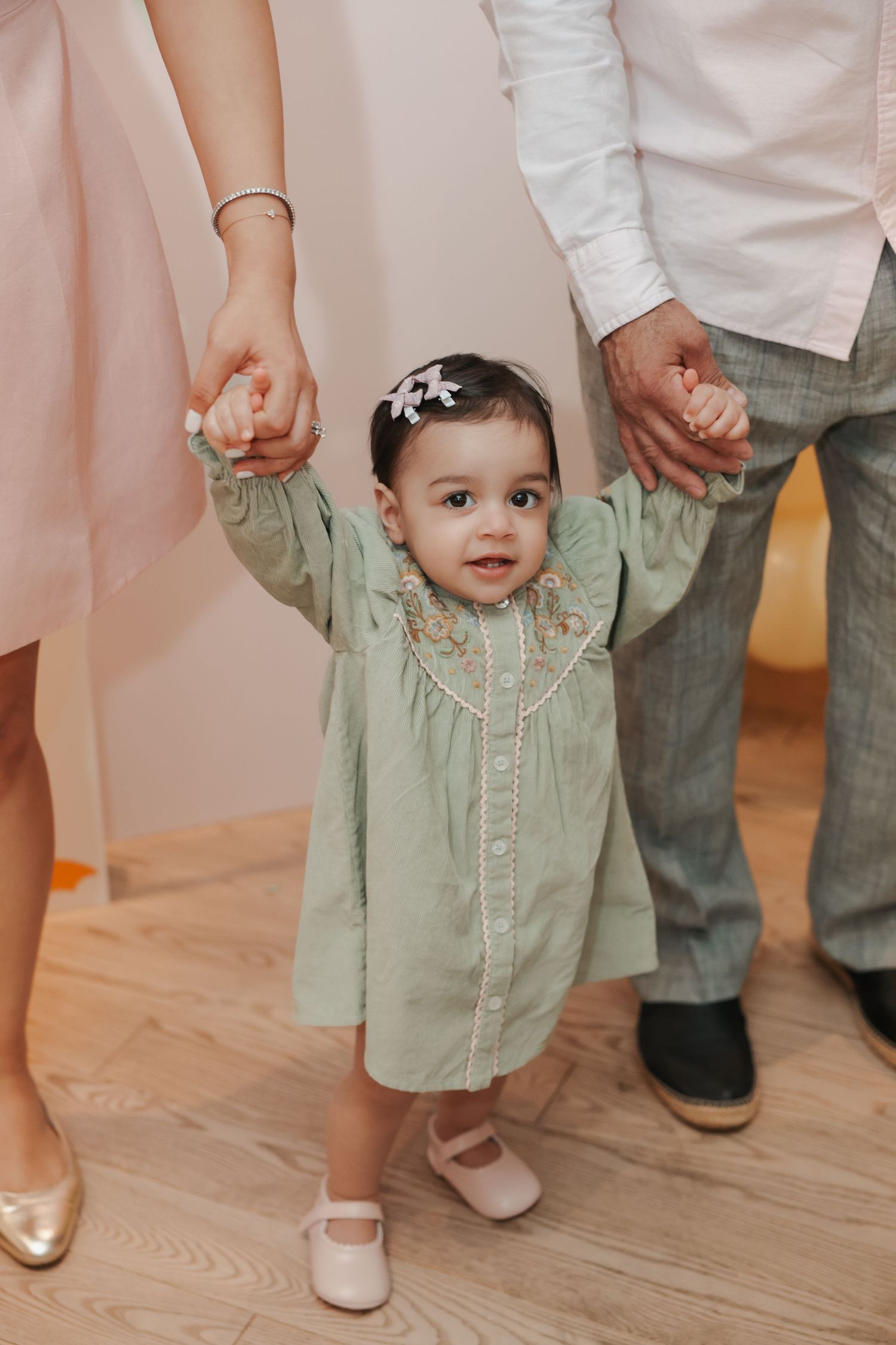 Toddler girl in green dress held by parents hands learning to walk on wood floor.