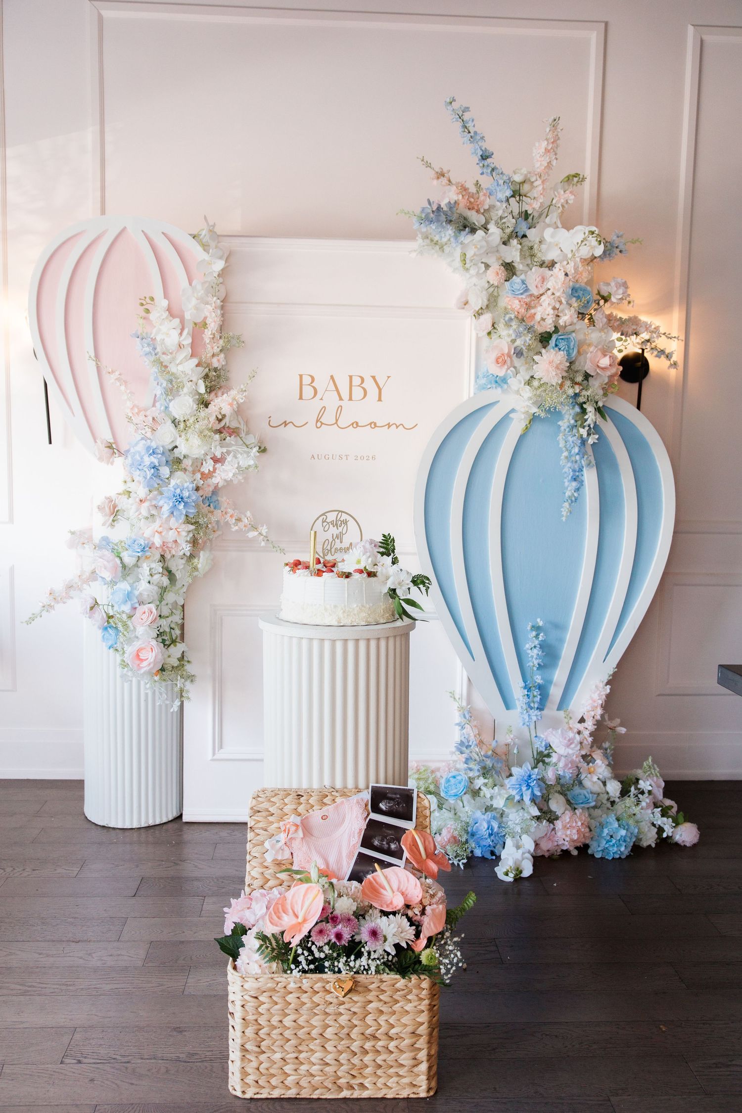 Baby in Bloom shower backdrop with pink and blue hot air balloons, white cake, and floral arrangements.