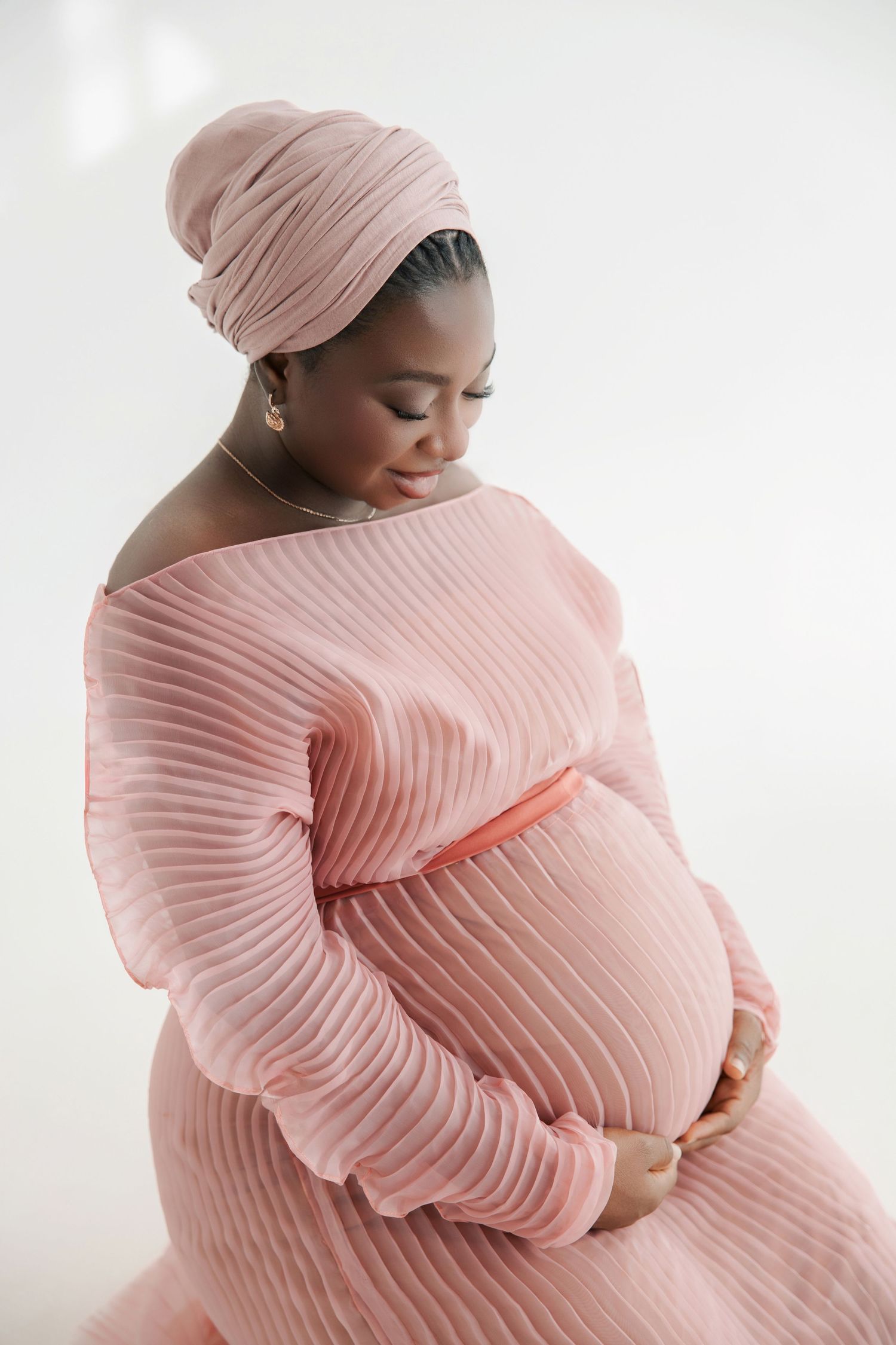 Pregnant woman in pink pleated dress and head wrap cradling baby bump against white background.