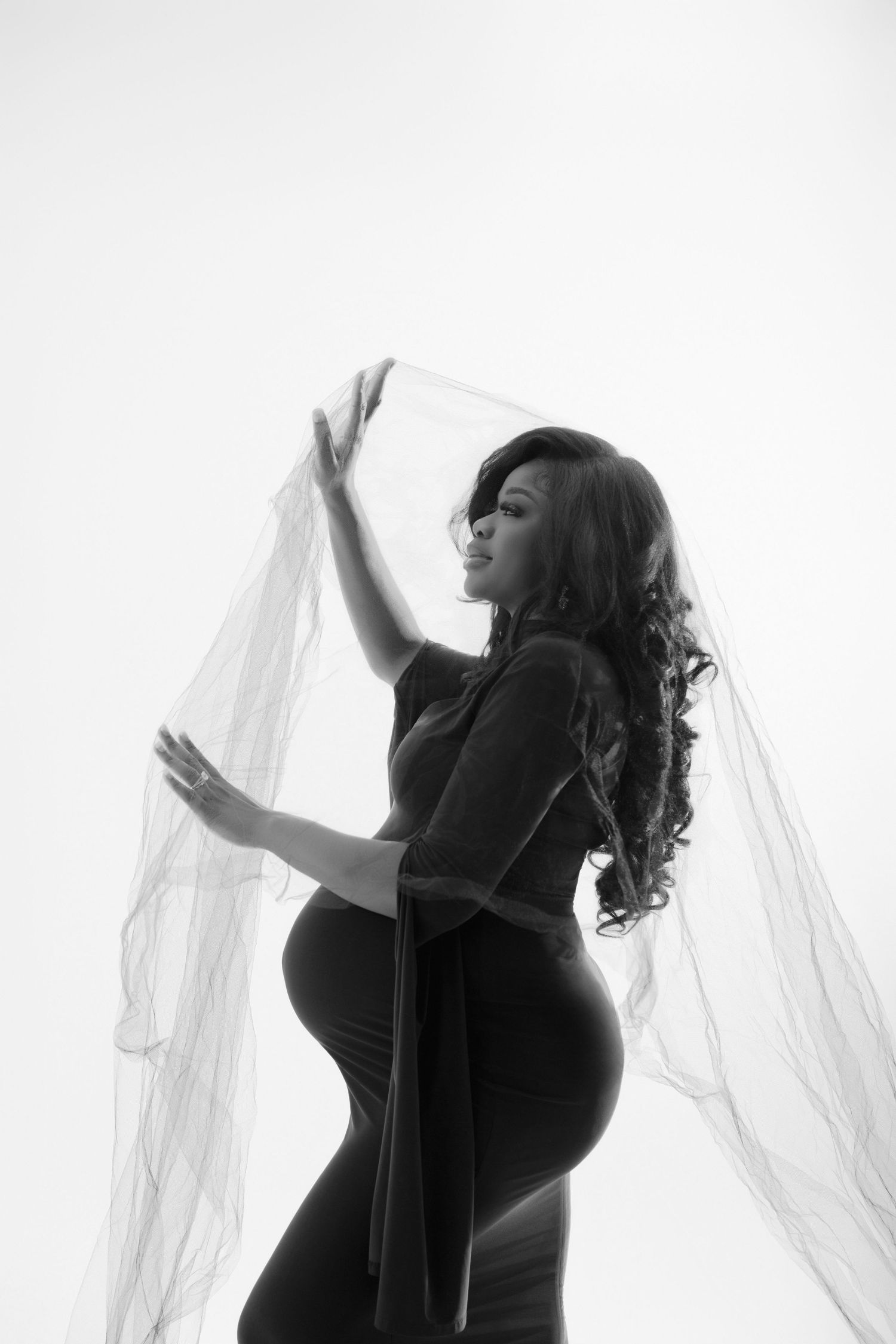Black and white maternity photo of pregnant woman in elegant dress holding sheer fabric, studio portrait.