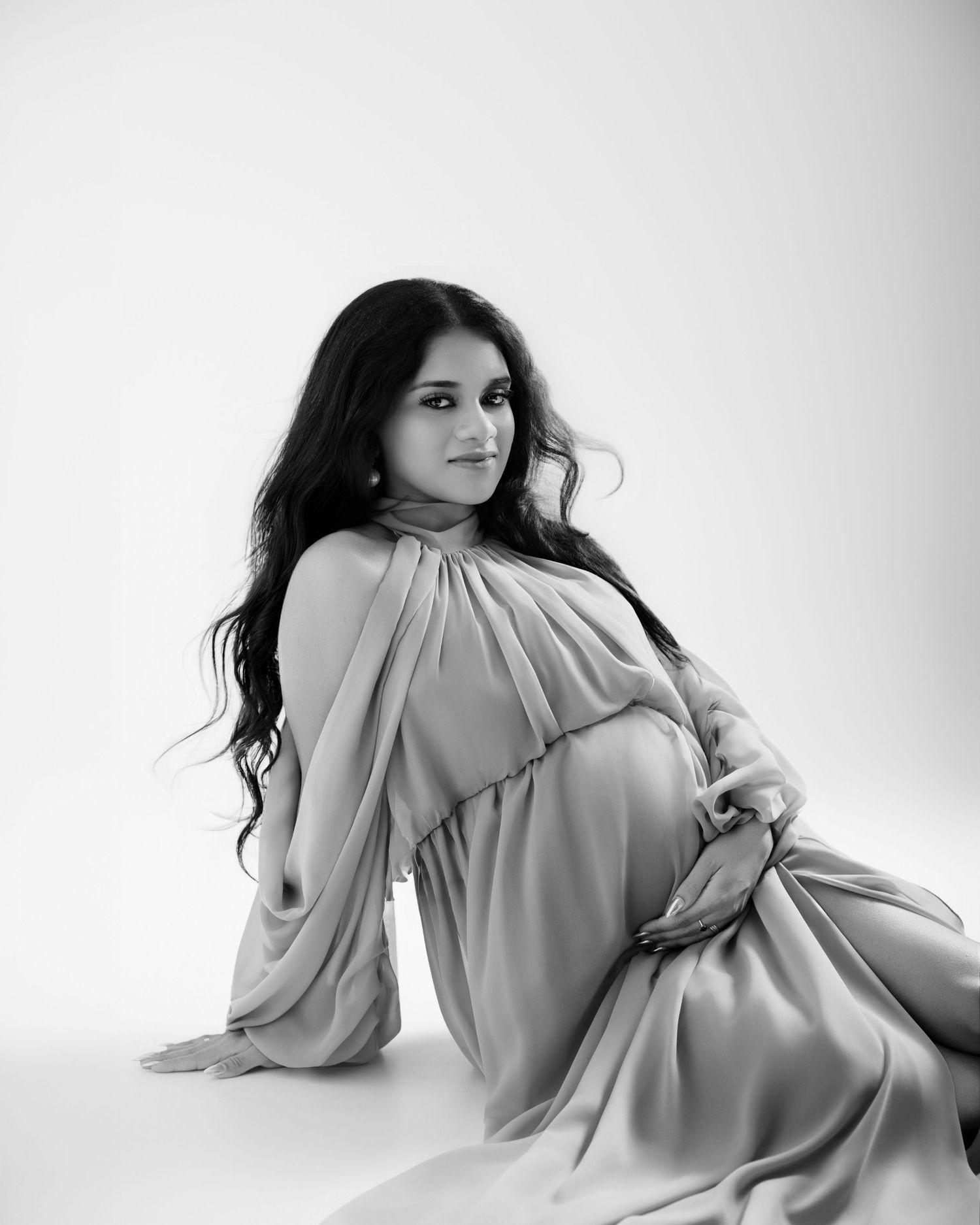 Black and white maternity photo of a woman in elegant draped gown sitting gracefully on white studio floor.
