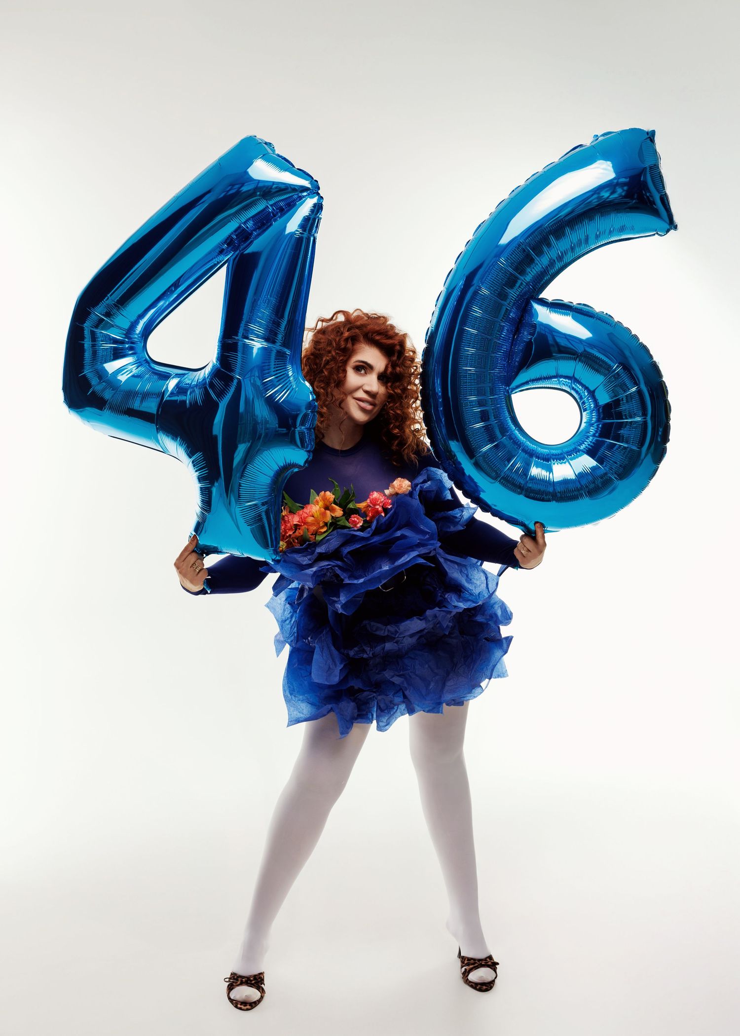 Joyful curly redhead woman in blue ruffled dress holding large blue number 46 balloons and flowers on white background.