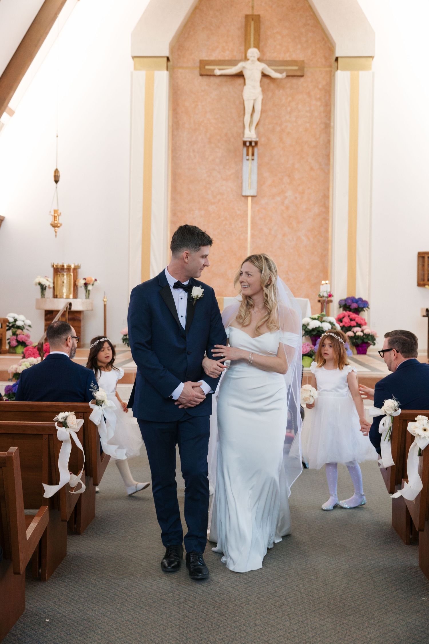 Bride and groom walk down church aisle after wedding ceremony, with flower girls and guests in wooden pews behind them.
