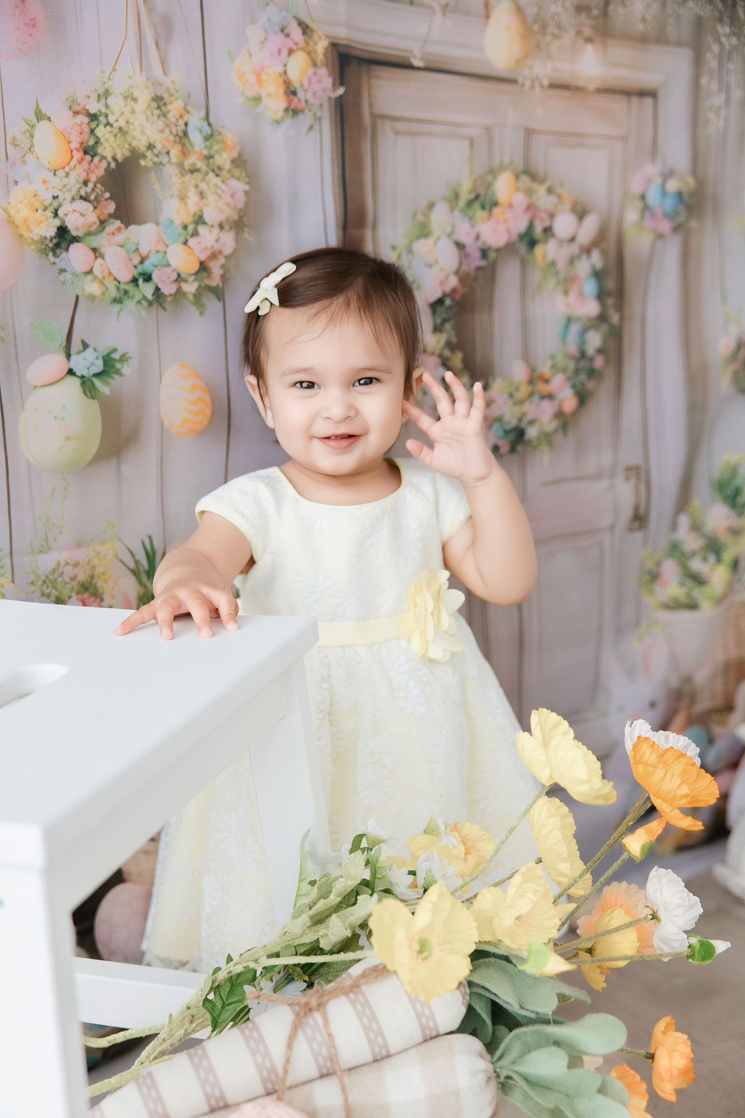 Smiling baby girl in white dress waving, surrounded by spring flowers and Easter wreaths in a pastel decorated studio.