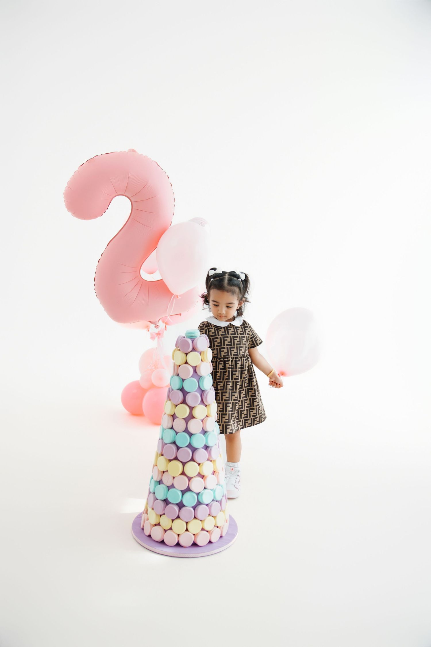 Toddler girl in designer dress stands beside a pastel macaron tower with a pink number 2 balloon at her 2nd birthday.