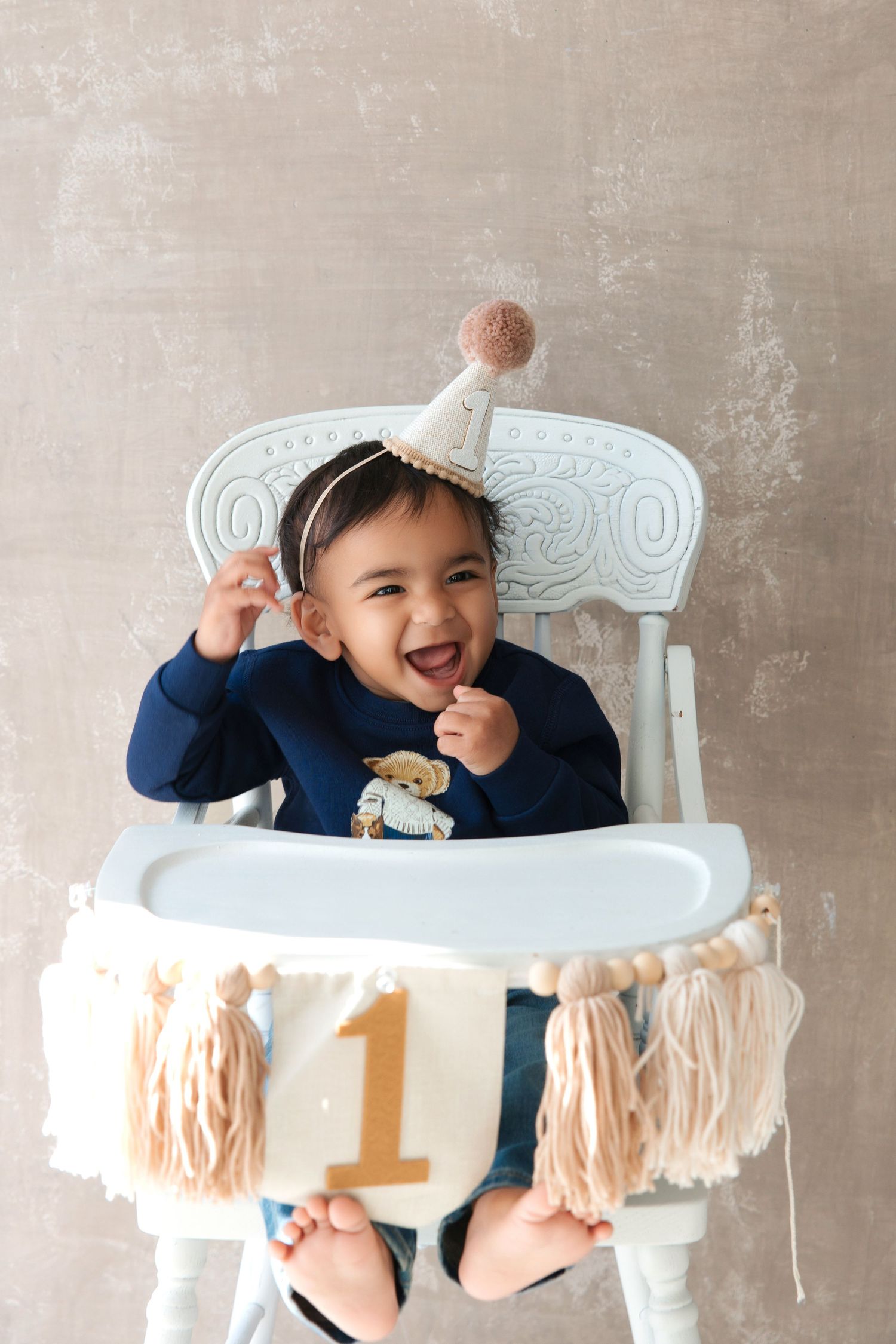 Happy baby in high chair wearing party hat celebrates first birthday with number 1 banner and tassels.