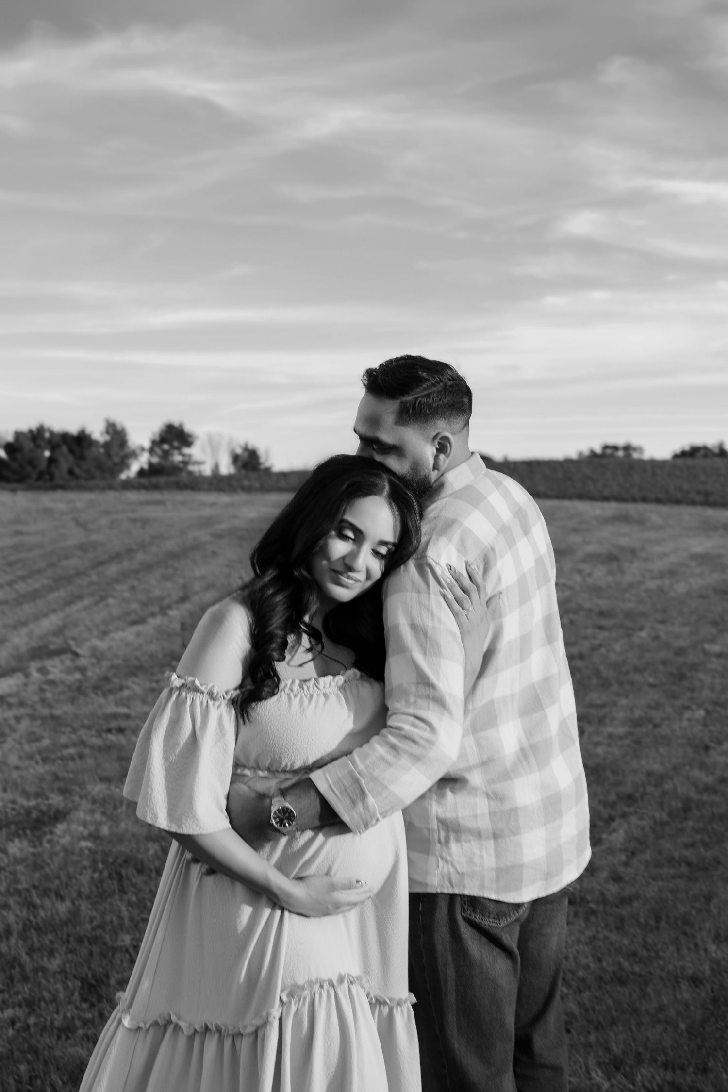 Black and white photo of a couple embracing in a field, woman in flowing dress cradling baby bump at sunset.