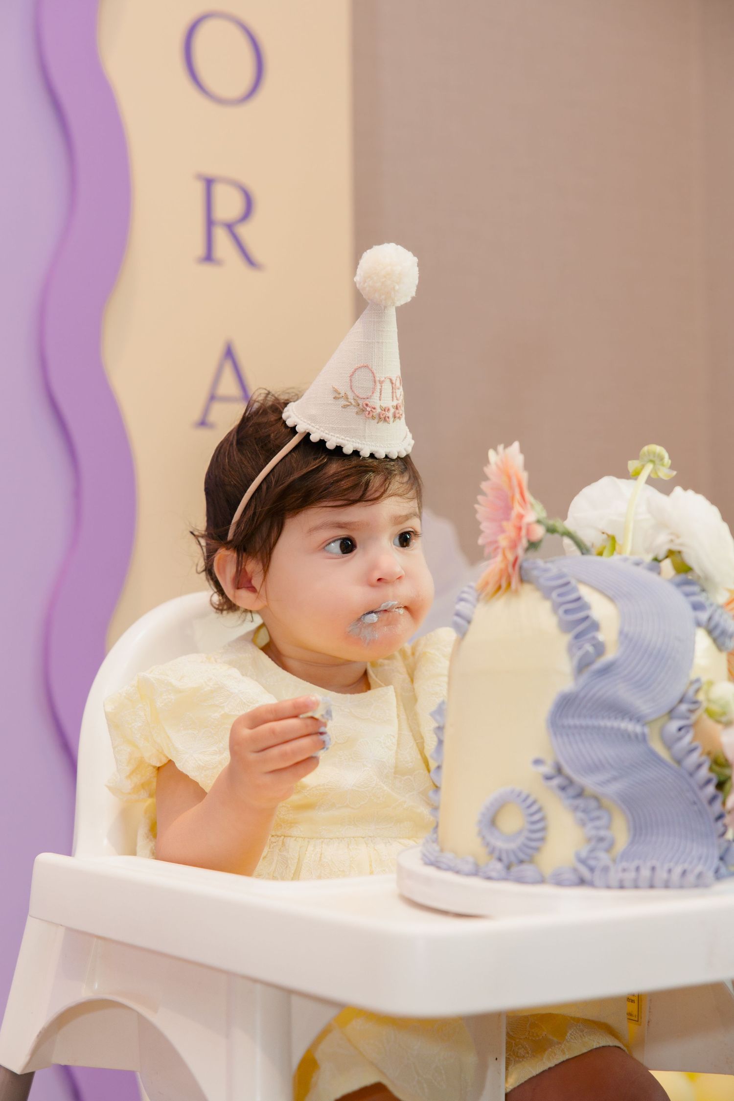 Baby girl in yellow dress and party hat sits in high chair next to first birthday cake.