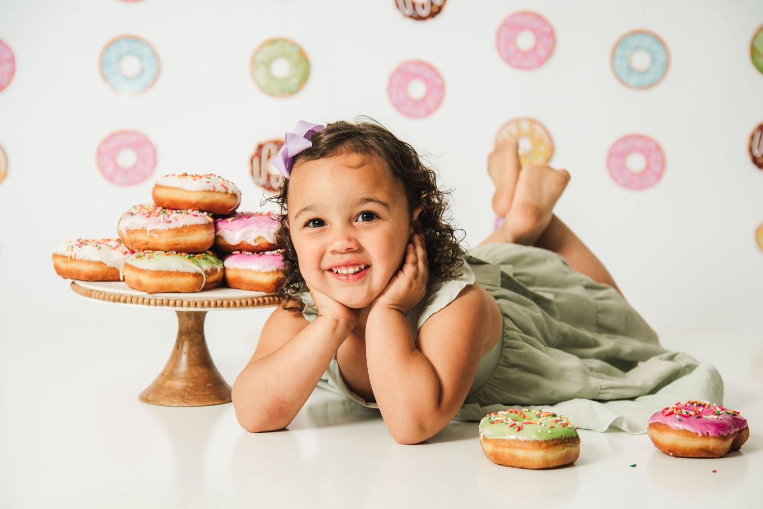 A child lies on the floor next to a cake stand filled with colorful frosted donuts against a donut-patterned backdrop.