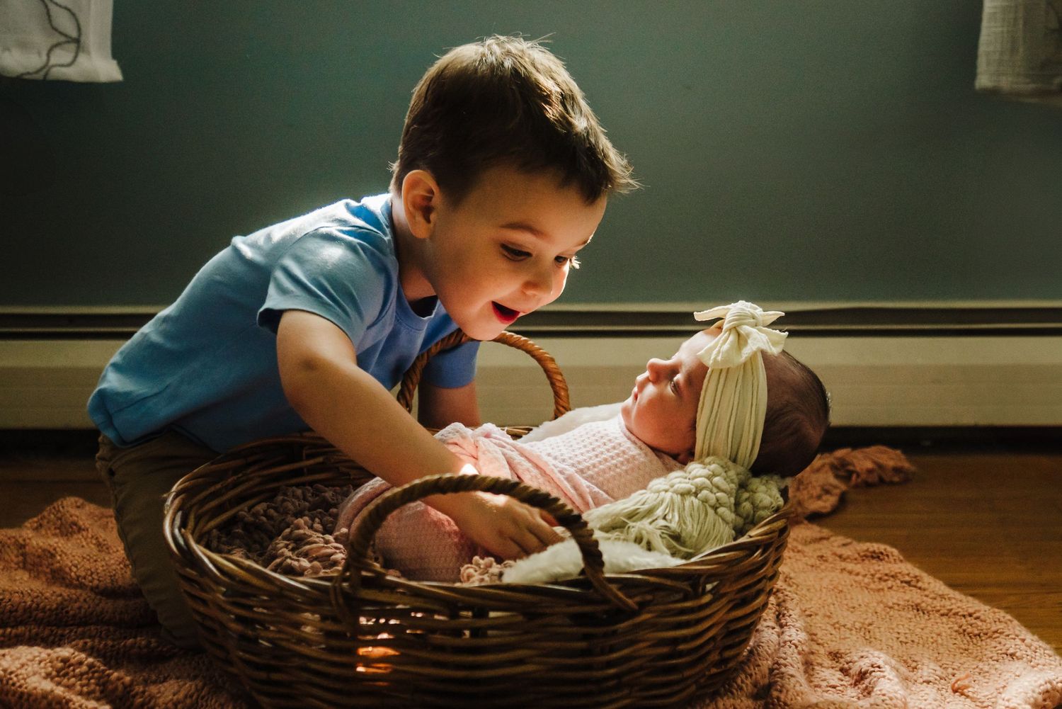 Young child lies on hardwood floor peering into a large woven basket in natural lighting.