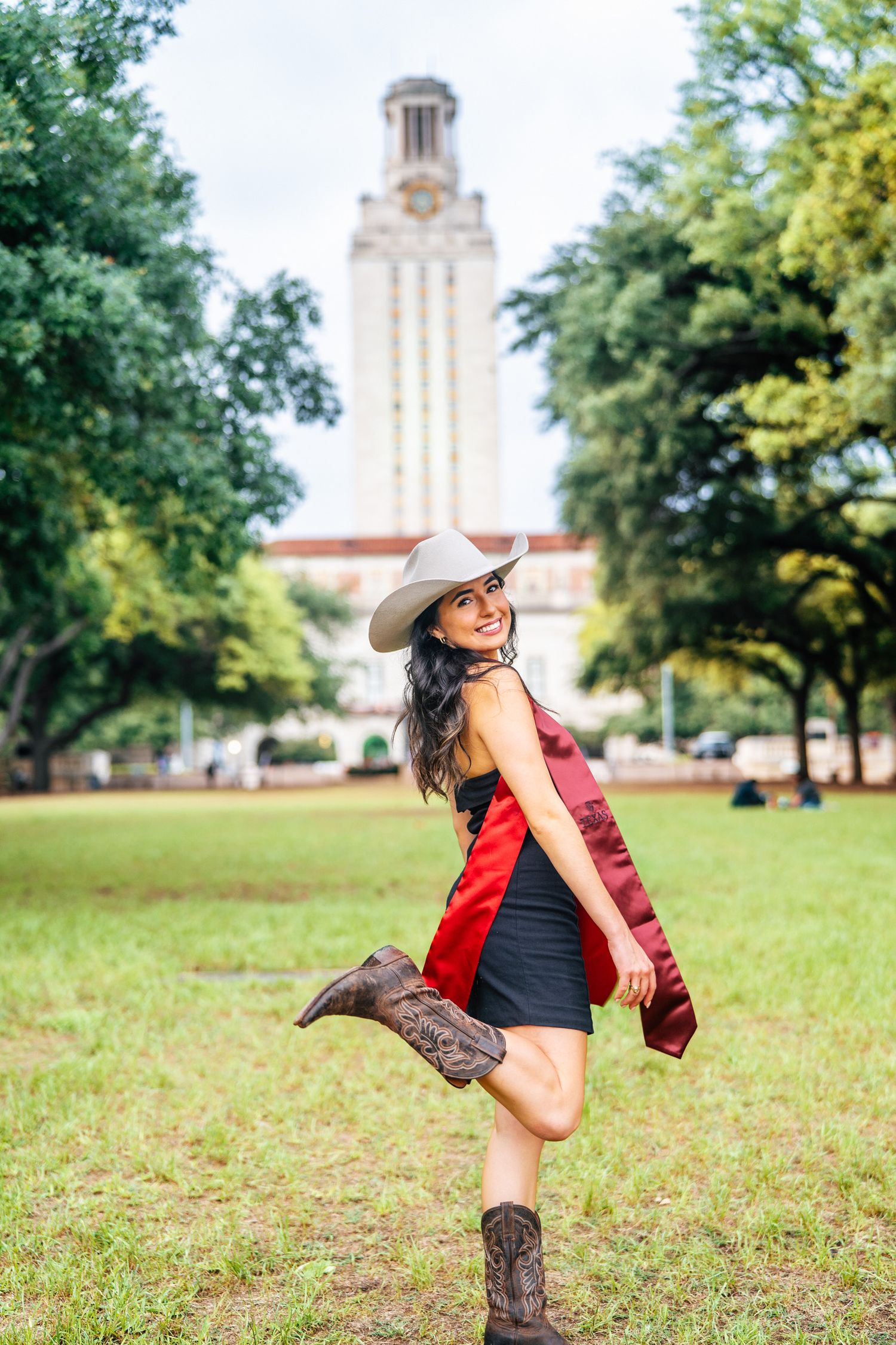 Playful graduate in cowboy boots strikes pose on lawn with UT Tower in background.