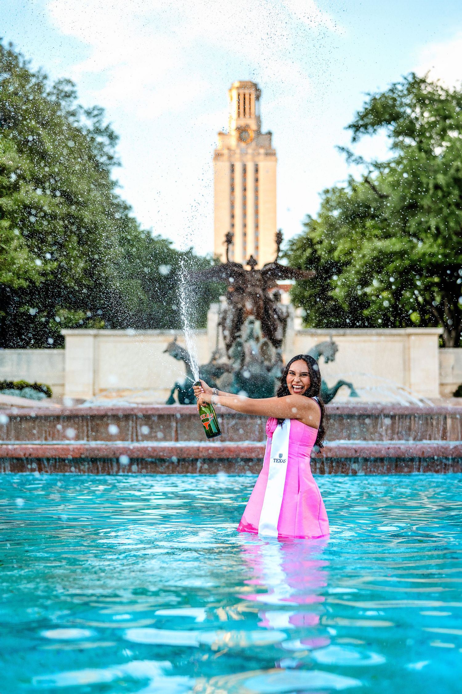 Graduate in pink dress celebrates in campus fountain with Tower and sculptures in background.