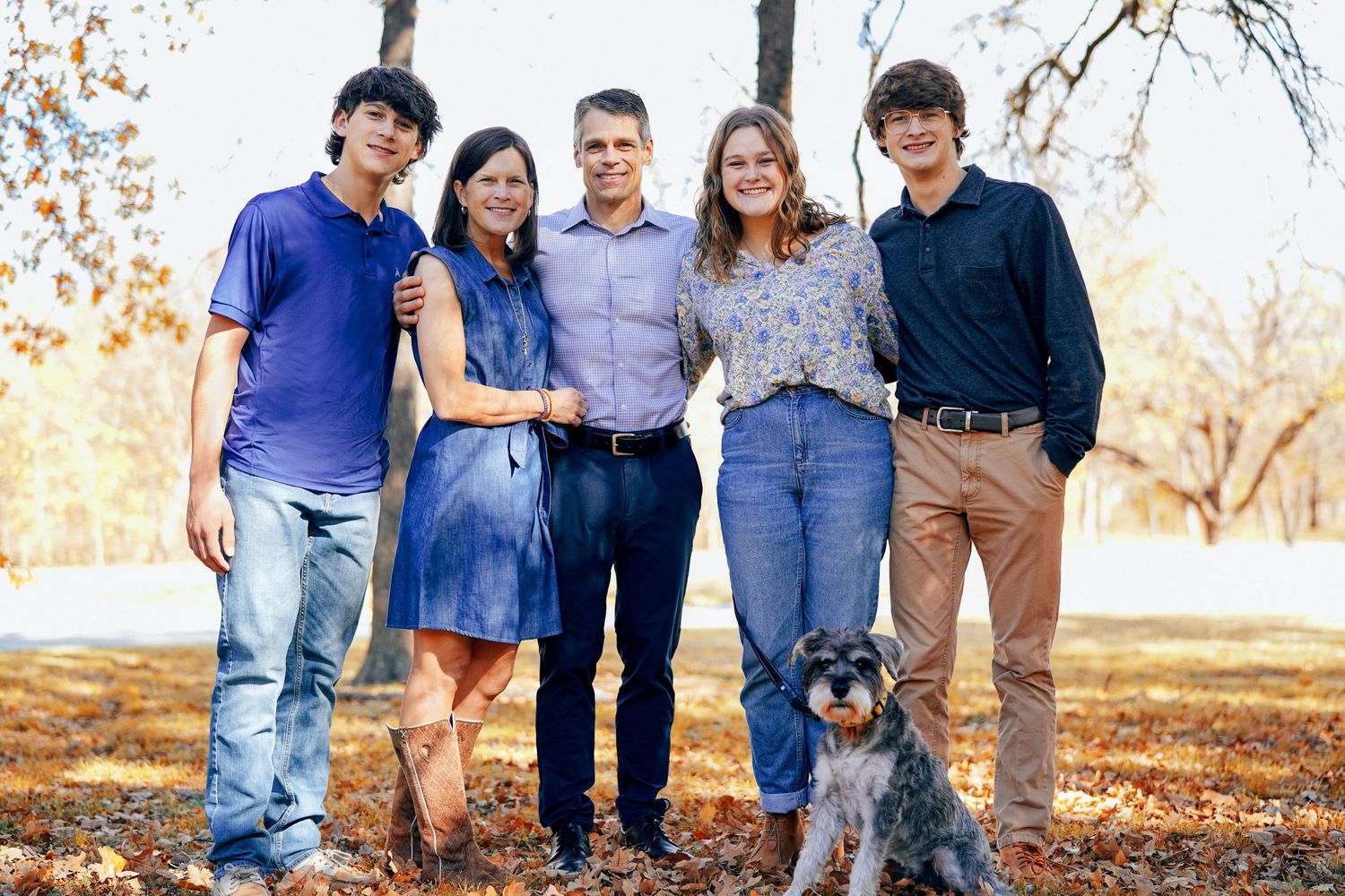 Family of five posing together with their dog on fallen leaves during autumn.