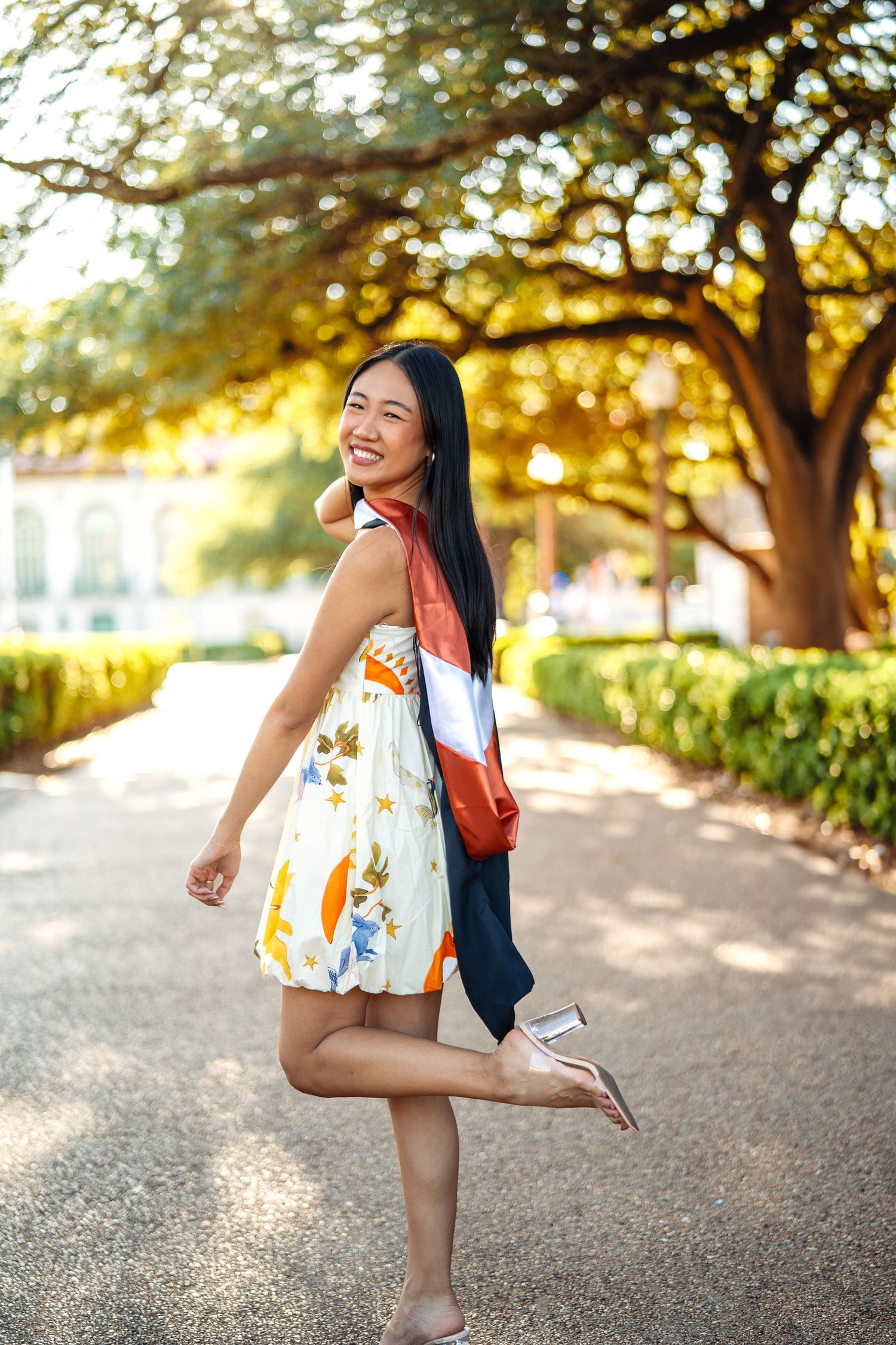 A person in a patterned sundress with orange bag strikes playful pose on tree-lined path.