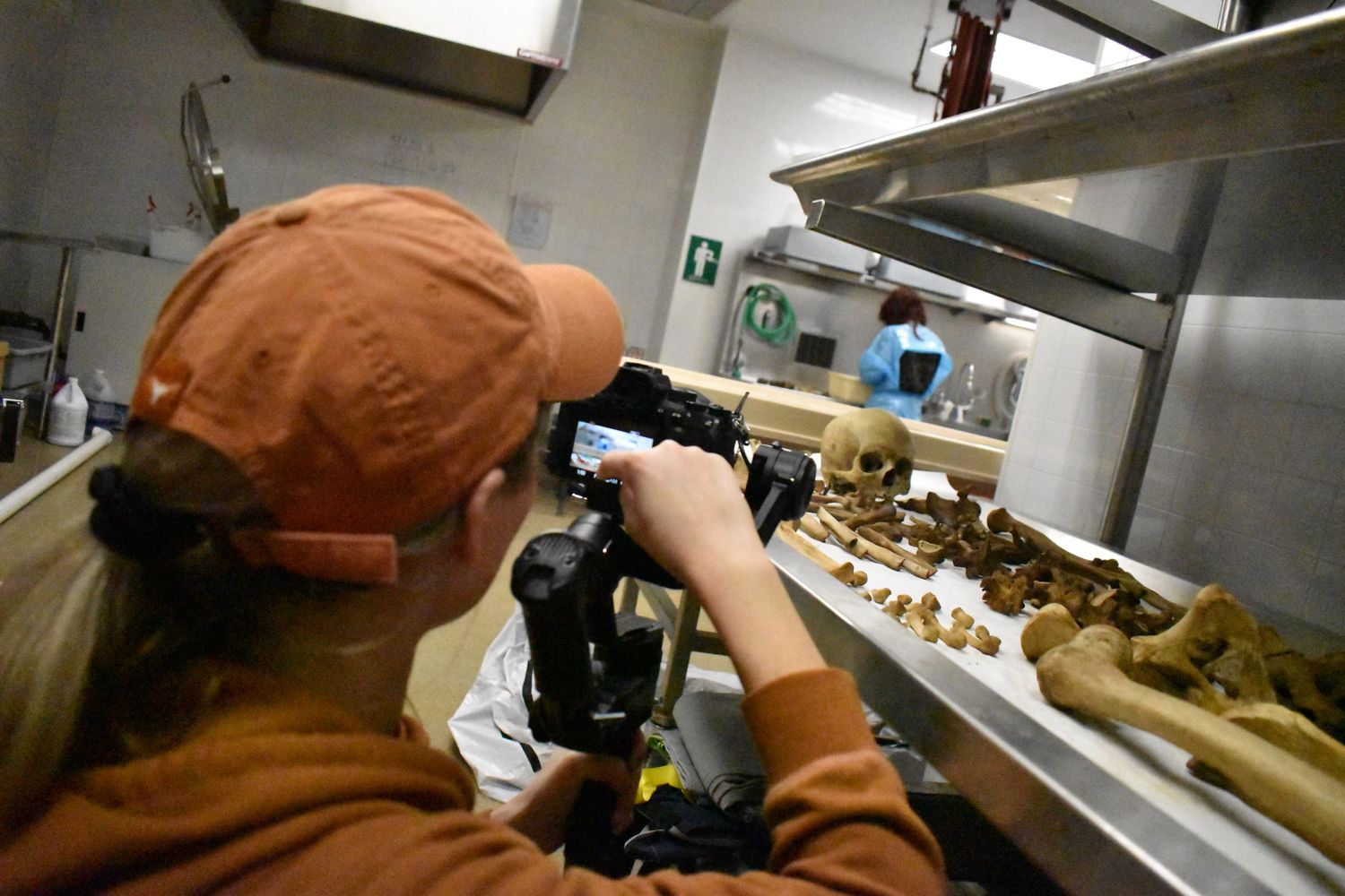 Camera operator wearing orange cap filming food preparation in commercial kitchen.
