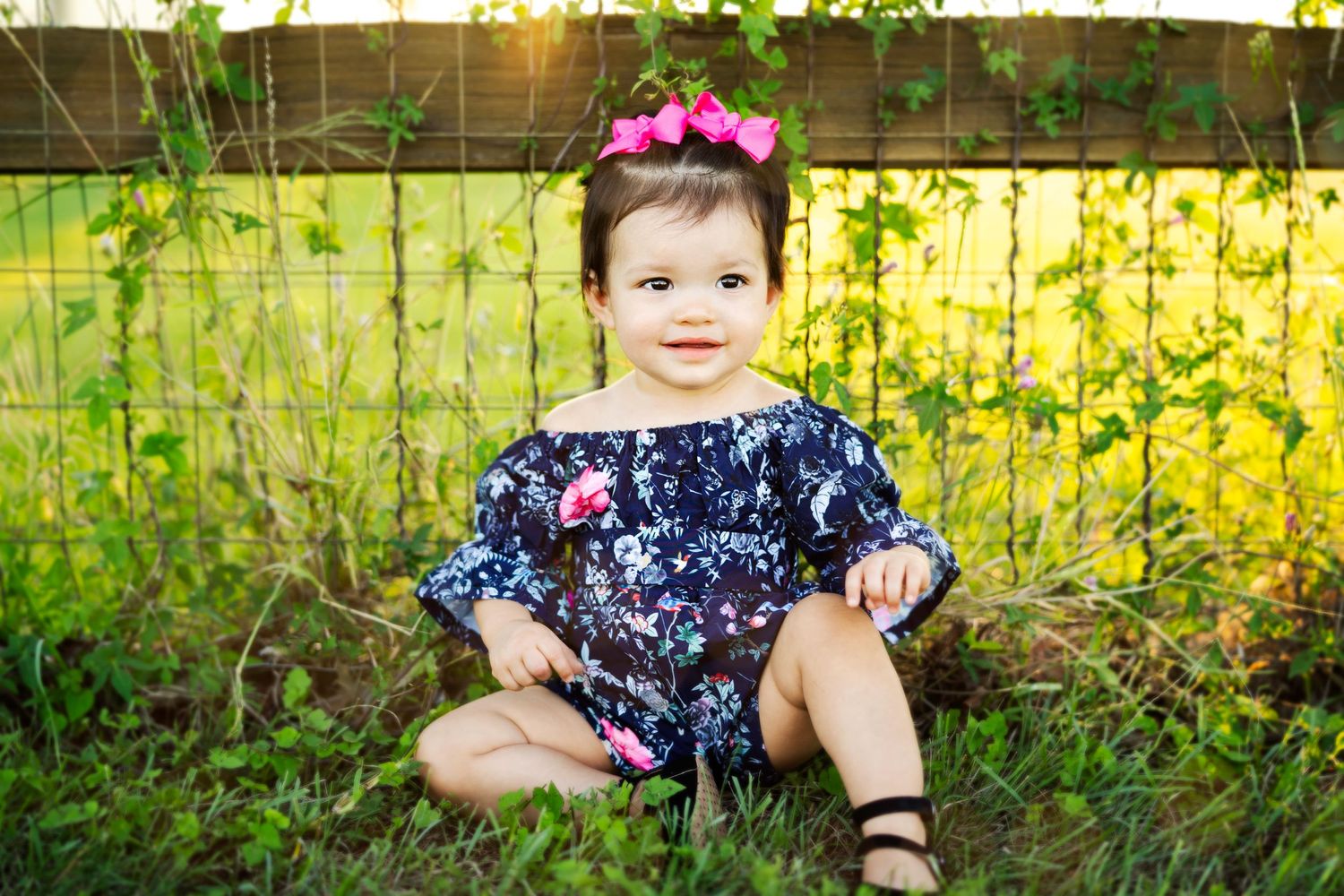 A child in navy floral dress and pink bow sits in grass during golden hour.