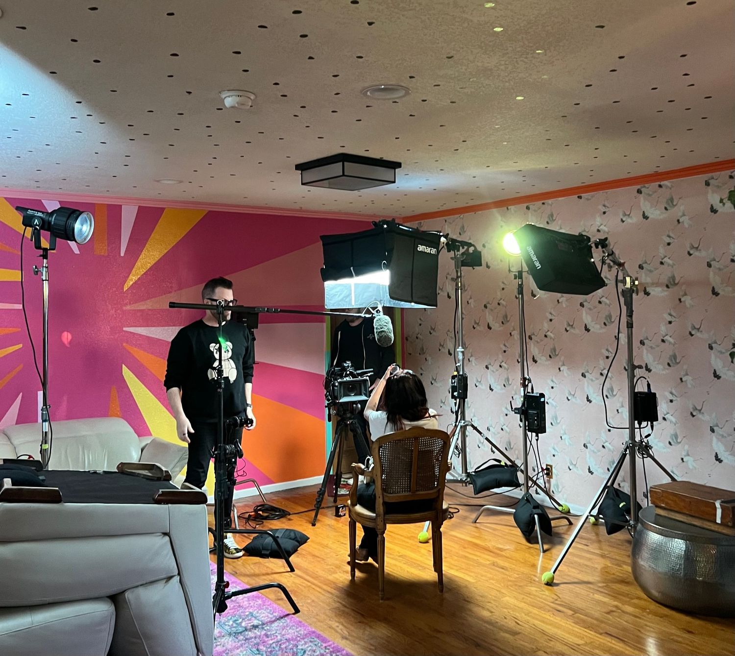 Film production setup in a colorful room with lighting equipment and cameras pointed at a seated subject.