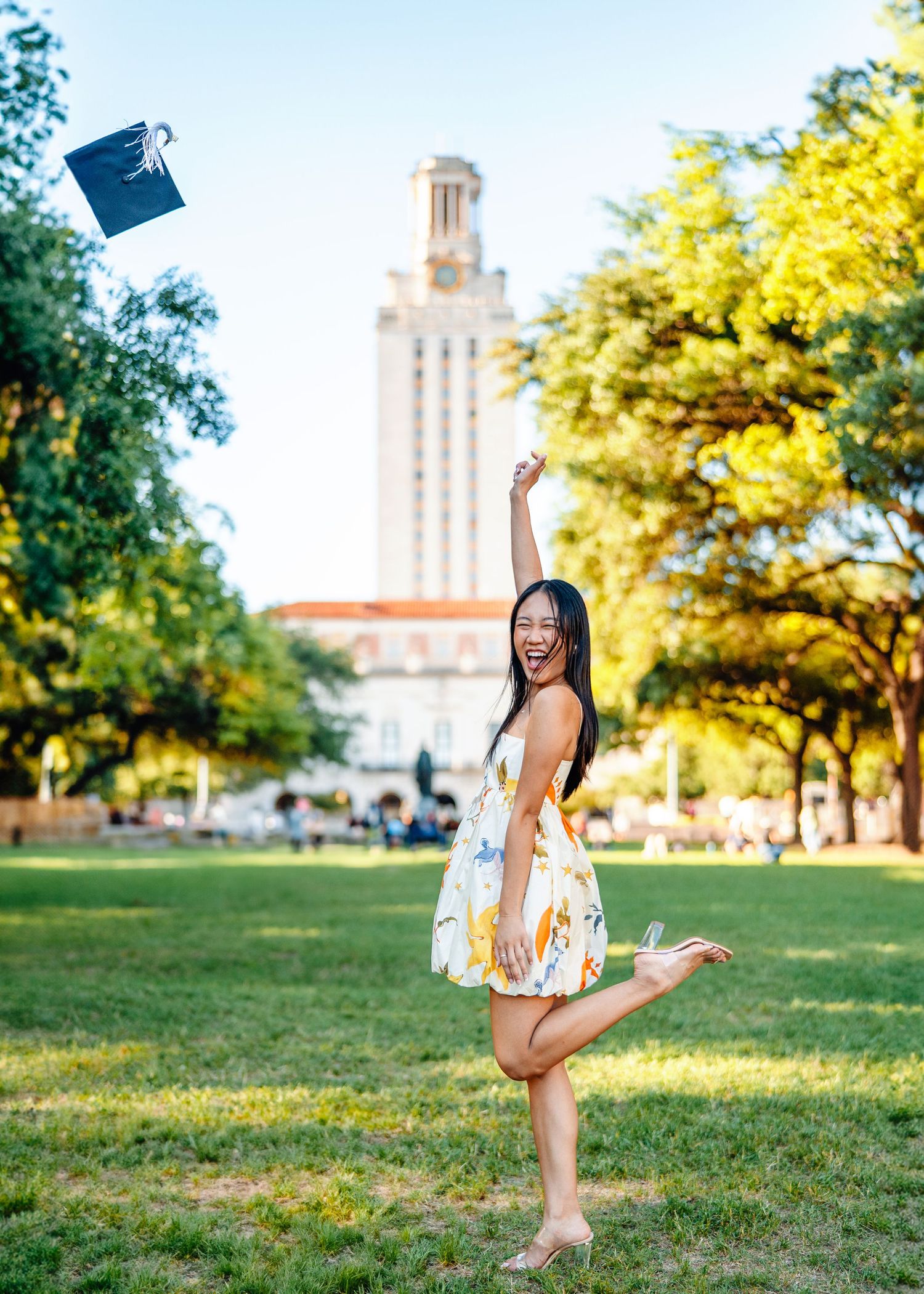 Graduate in floral dress throws graduation cap on sunny university campus lawn with iconic tower in background.