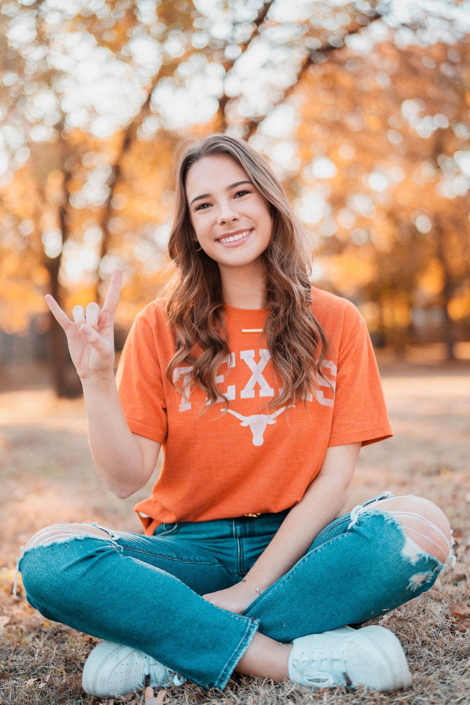 Person sitting on ground wearing orange shirt and teal jeans during autumn photoshoot.