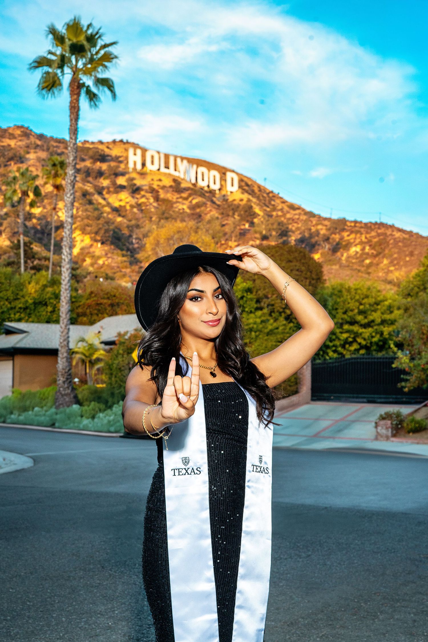 UTLA Student wearing black hat and graduation stole poses in front of iconic Hollywood sign with palm tree in Los Angeles.