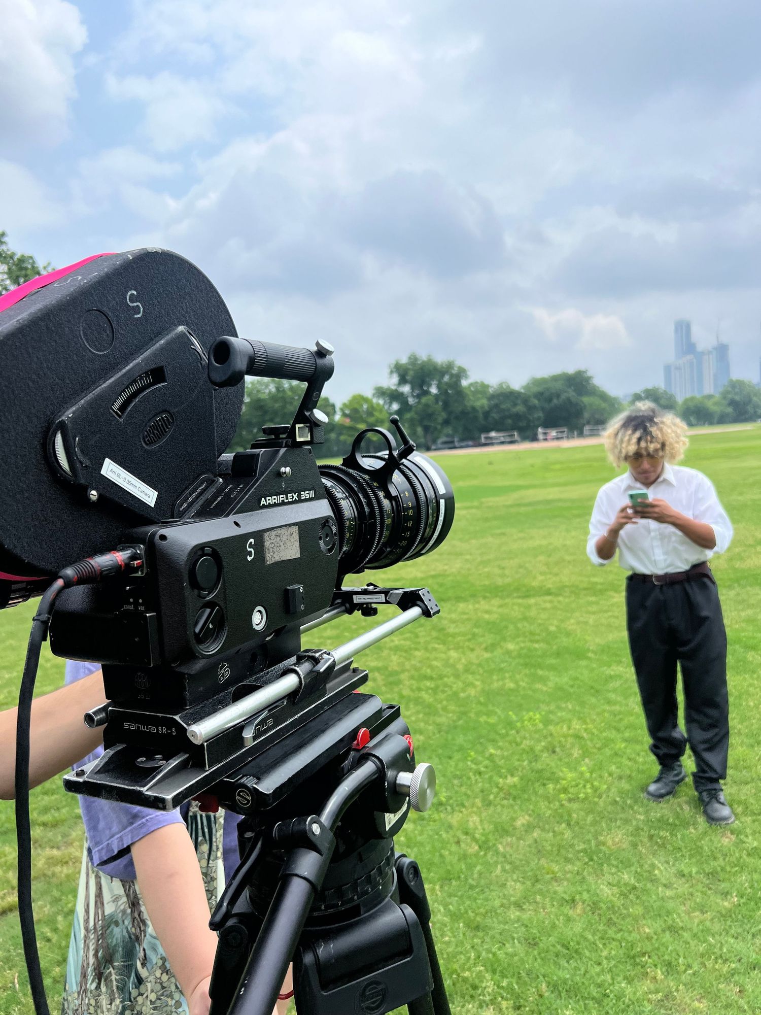 Professional camera operator films a subject on green grass with city skyline in background.