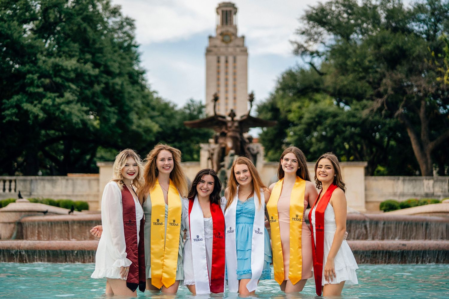 Group of graduates in vibrant dresses pose by fountain with iconic UT Austin tower in background.