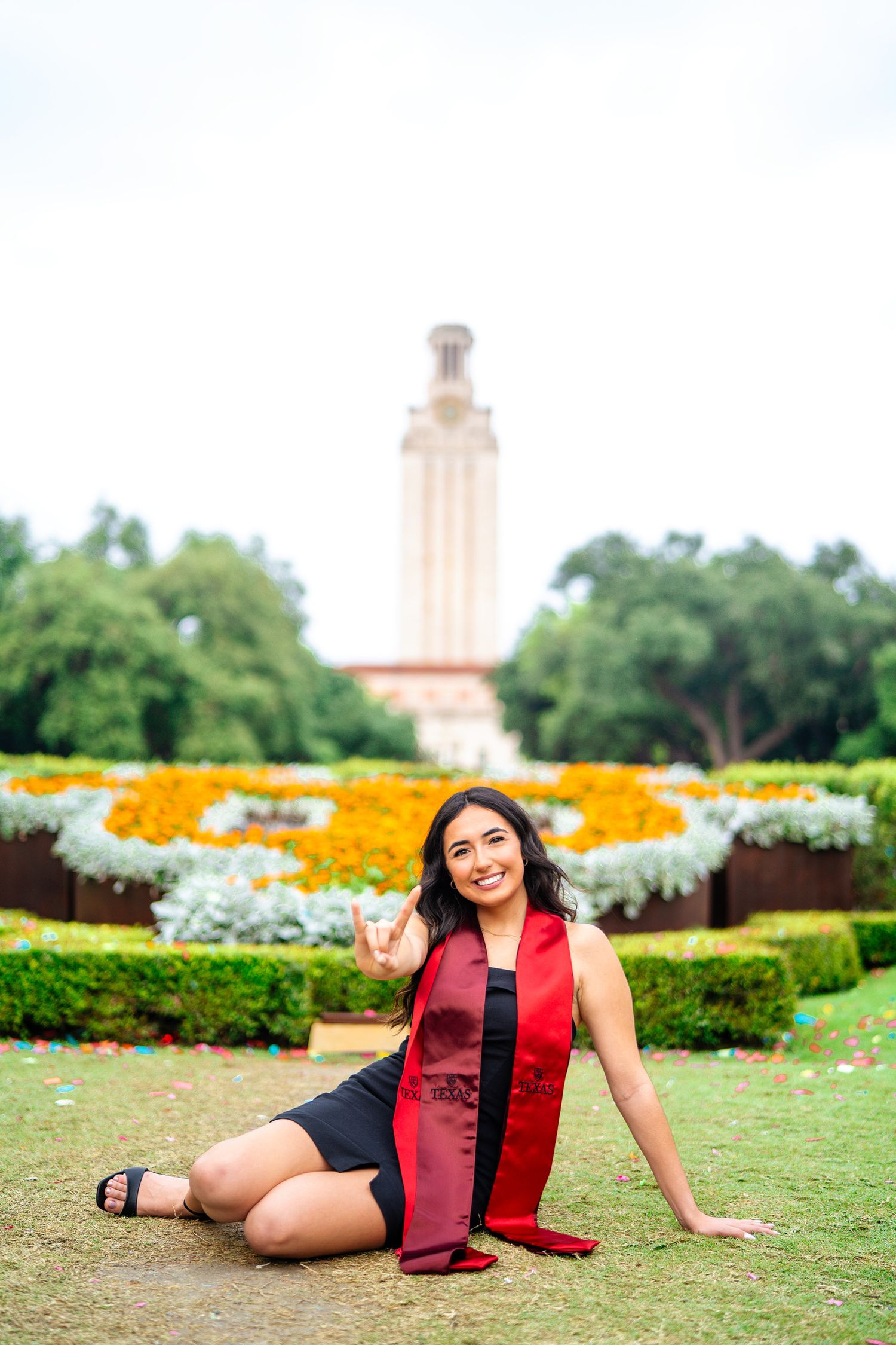 Graduate in red stole poses in garden with UT Tower and yellow wildflowers in background.