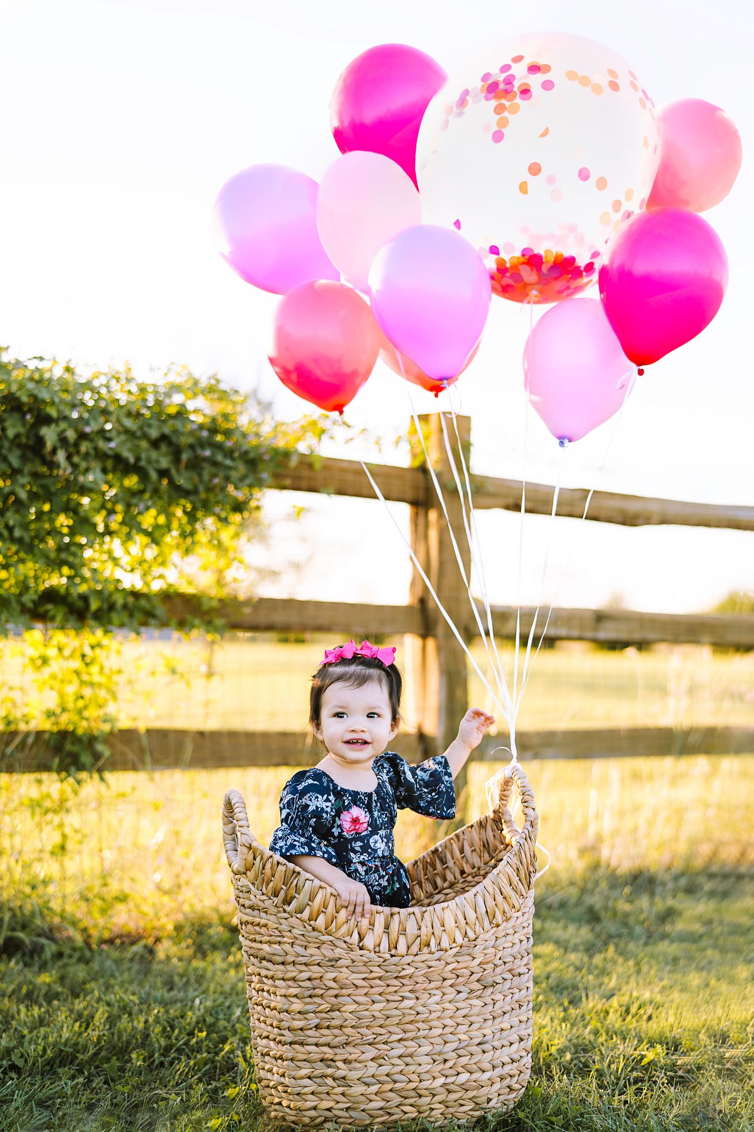 A child sits in woven basket with pink and white balloons during sunset.